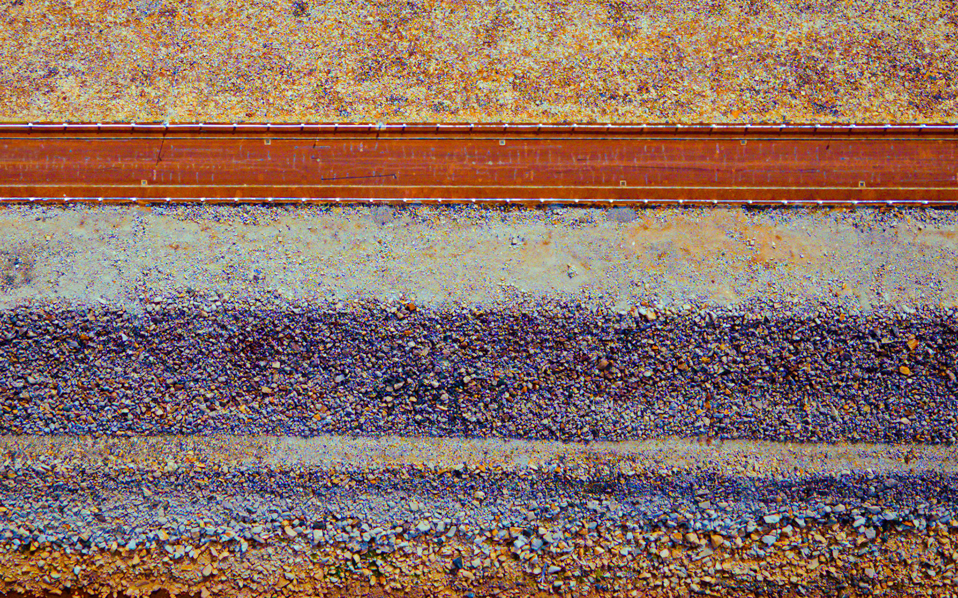 A wide view shows a railroad track cutting through land with different shades of brown and gray. The track is straight and leads far into the distance under clear skies.