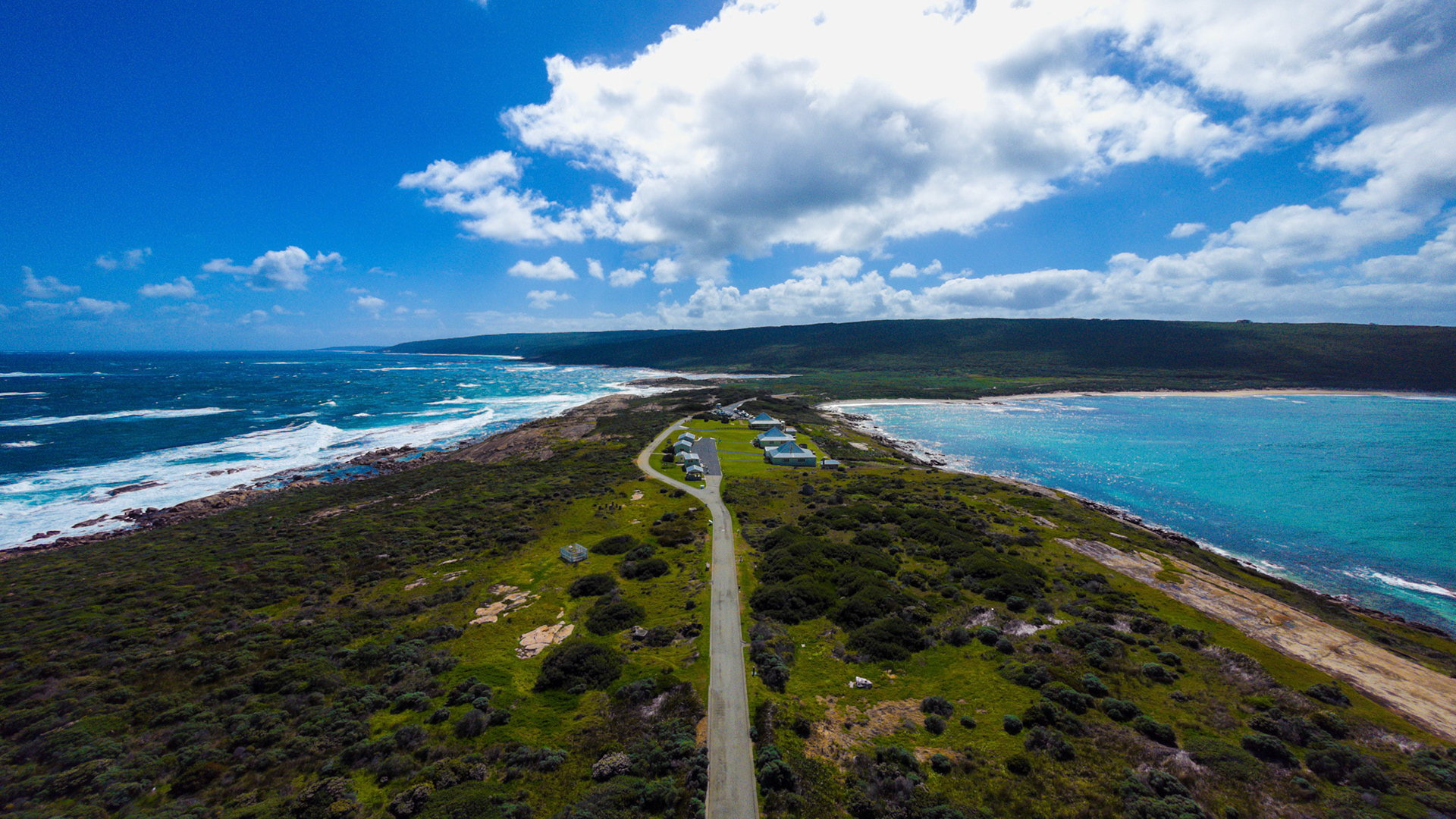 A stunning aerial view captures a winding road leading to a cluster of seaside houses on a lush cliffside. The vibrant blue ocean meets the rocky shoreline, while scattered clouds decorate the clear sky, creating a picturesque coastal scene.