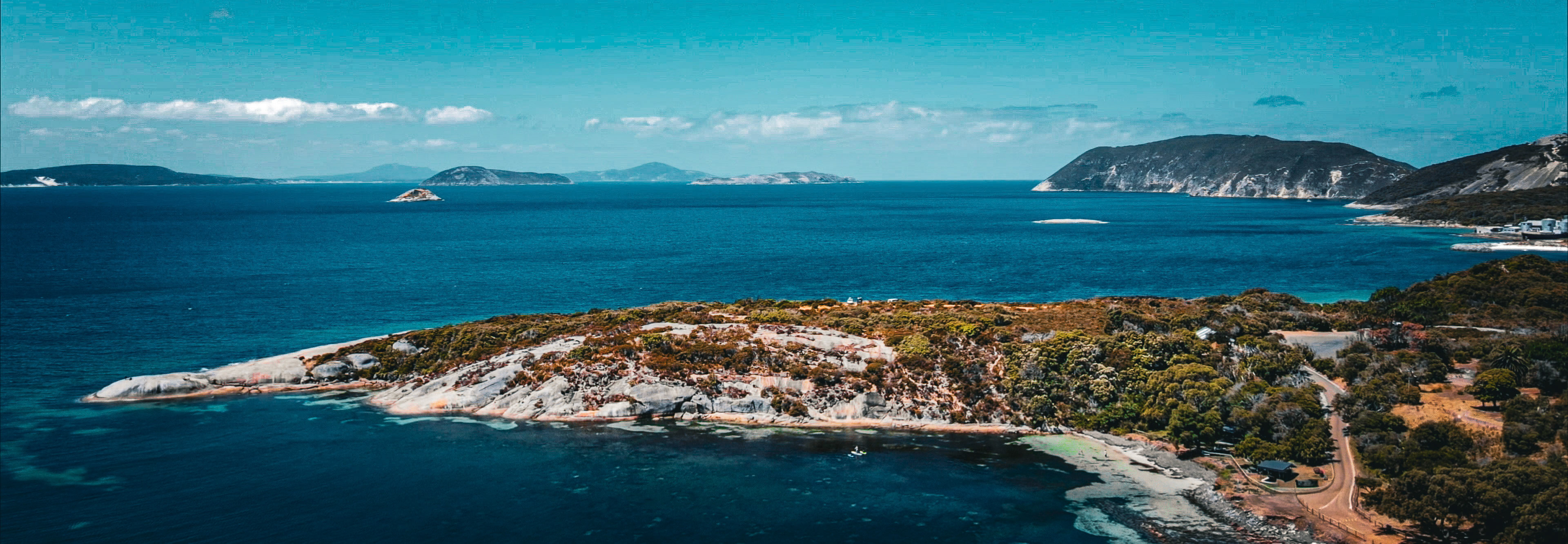 A panoramic aerial view of a rugged coastline where white granite outcrops and dense coastal scrub meet brilliant turquoise water. A winding road skirts a sheltered cove in the foreground, while small islands and distant, hazy hills rise from a deep blue sea under a bright sky dotted with light clouds.