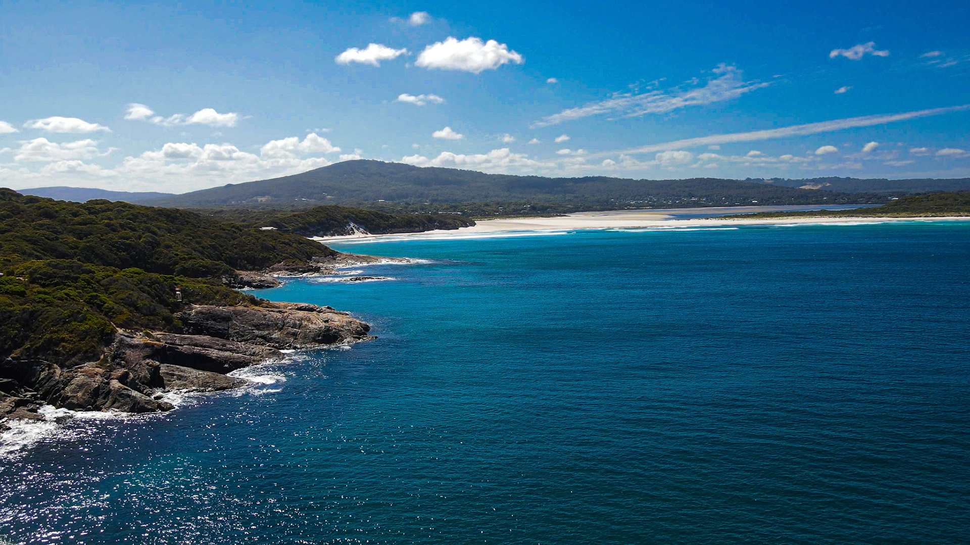 An expansive aerial view showcases a coastal landscape with turquoise ocean waters, sandy beach, and lush green hills under a bright sky.