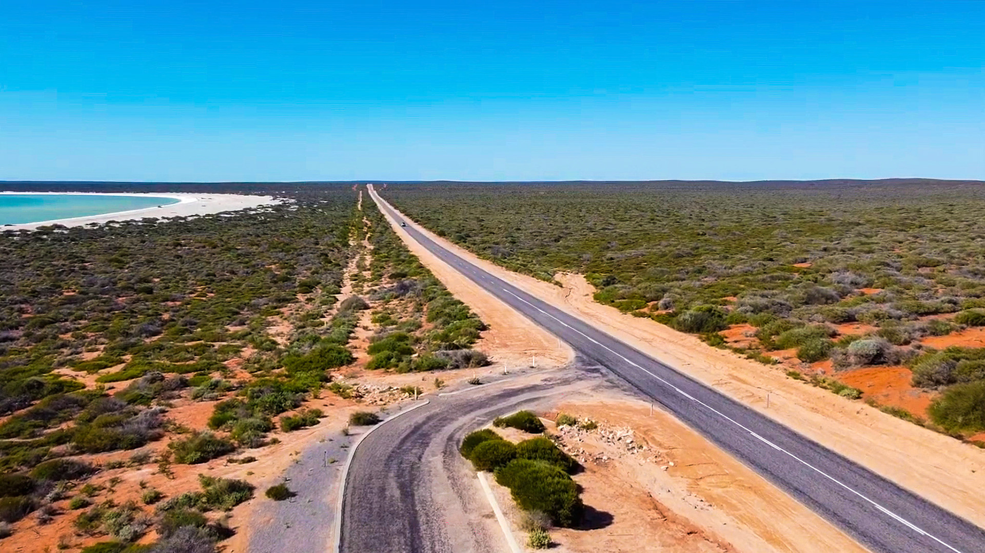 An aerial view shows a long, empty highway leading through a vast desert landscape towards a distant coastline and turquoise ocean.