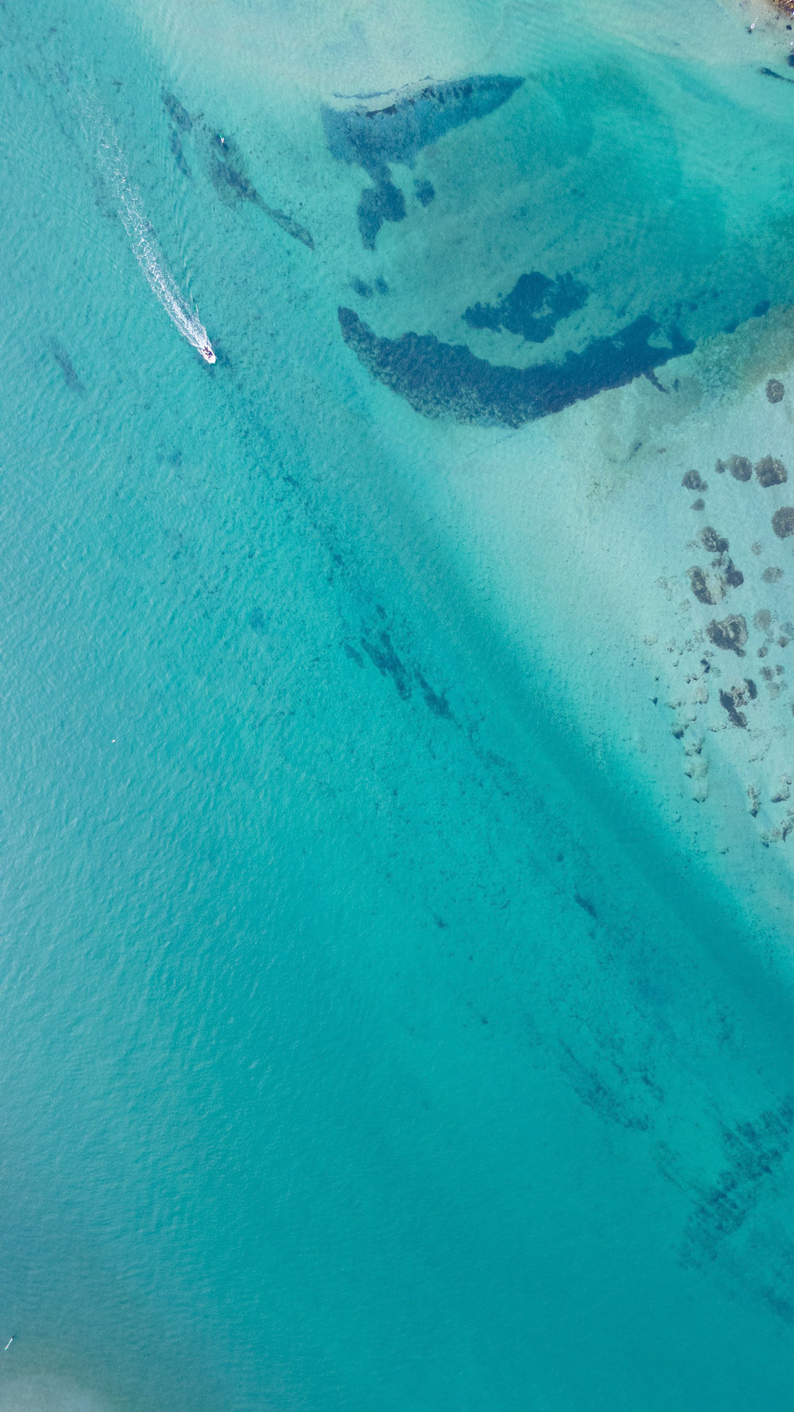 Immerse yourself in this stunning aerial shot capturing the crystal-clear turquoise waters of a tropical paradise, with a small boat gently gliding across the surface. The tranquil water reflects varying shades of blue and hints of underwater features, while the subtle texture of the waves adds depth to the landscape. This image exudes relaxation and captures the essence of a perfect day by the sea, ideal for travel and lifestyle projects.