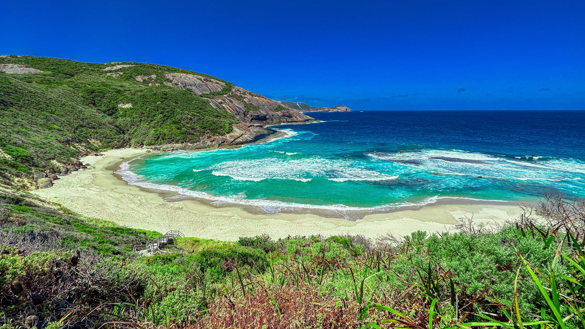 A tranquil coastal landscape features a secluded beach with turquoise waters, white waves, and vibrant blue skies, showcasing nature's beauty.