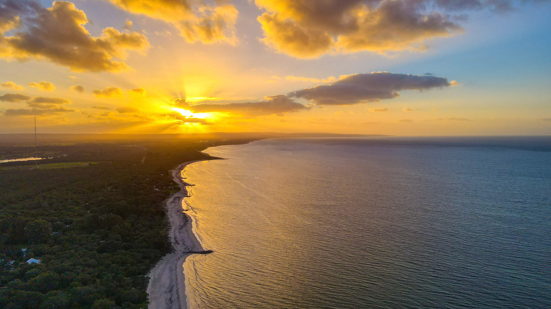 A breathtaking aerial view captures a stunning sunrise over a tranquil coastline. The golden sun beams through scattered clouds, casting warm hues over the serene ocean waves and sandy beach. This peaceful scene showcases the beauty of nature at dawn, perfect for concepts of tranquility and natural beauty.