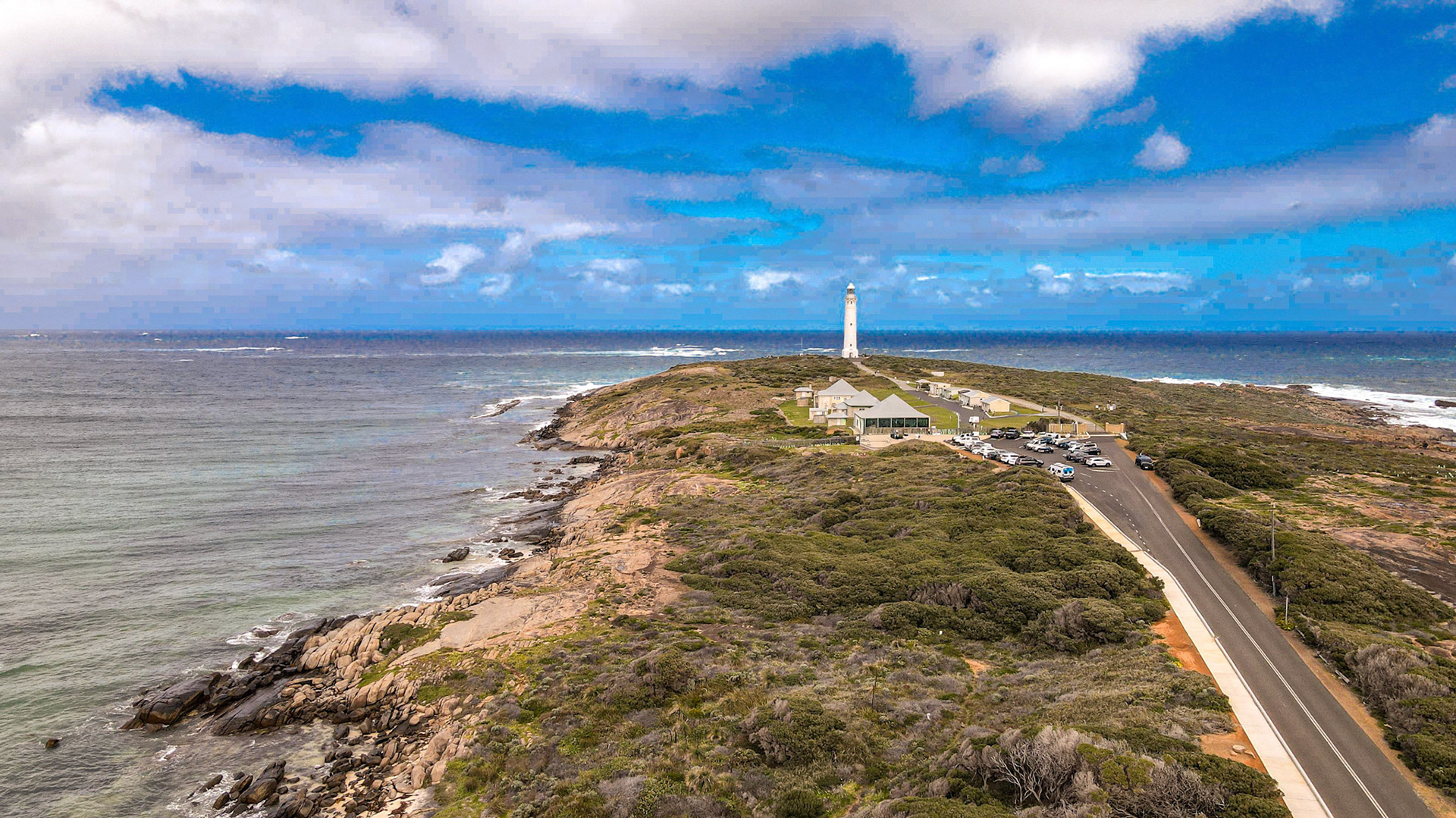 A stunning aerial view of a rocky shoreline leading to a prominent lighthouse. The scene captures the lush greenery of the coastal landscape alongside a winding road that guides visitors to this picturesque location. The vast expanse of the ocean stretches to the horizon under a partly cloudy sky, adding to the serene and tranquil ambiance of the scene.