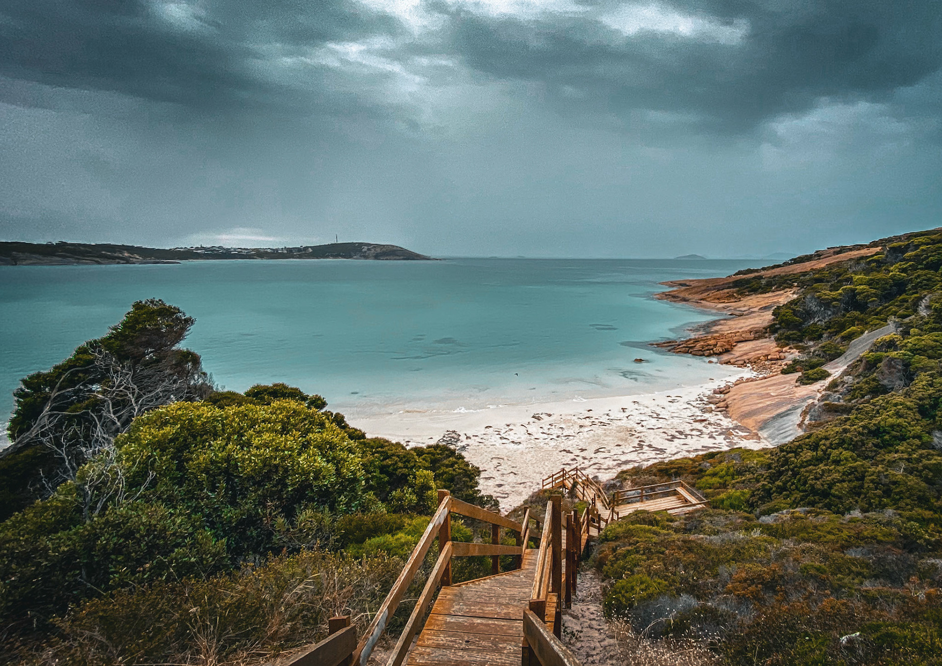 A wooden staircase leads to a secluded beach, surrounded by lush vegetation and rocky shores, evoking tranquility and adventure.