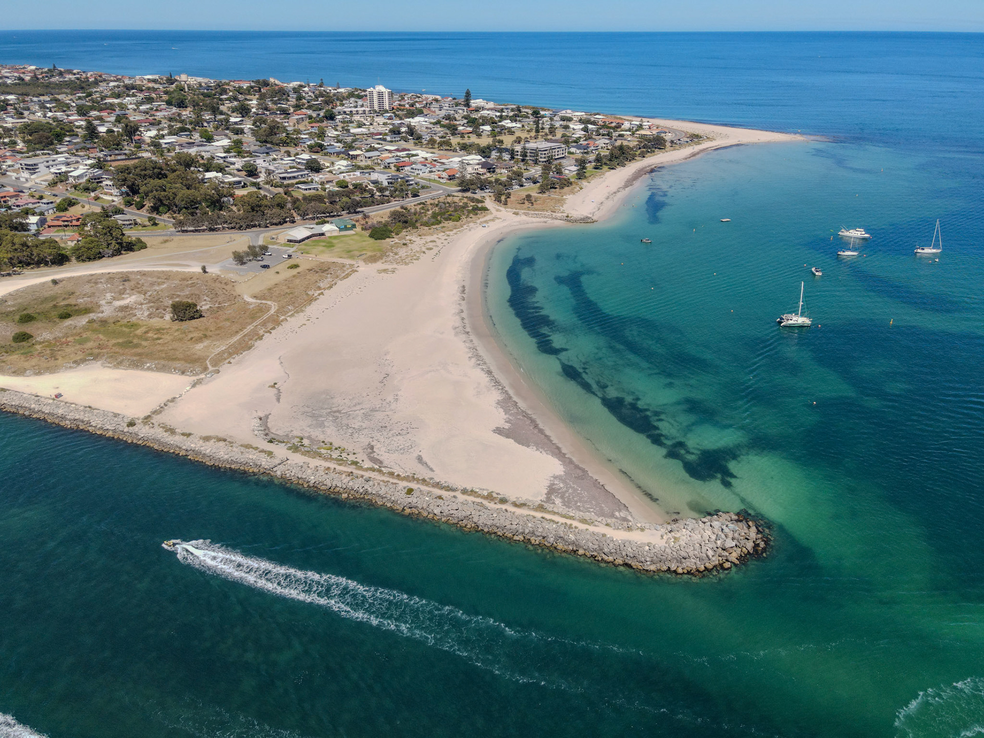 A high-angle aerial view of a crescent-shaped sandspit forming a calm lagoon. Clear turquoise water with dark seagrass patches surrounds anchored sailboats and catamarans. A rock groyne protects the inlet while a speedboat cuts a white wake along the open water. Low-rise coastal neighborhoods and a few apartment buildings line the shoreline under a bright blue sky.