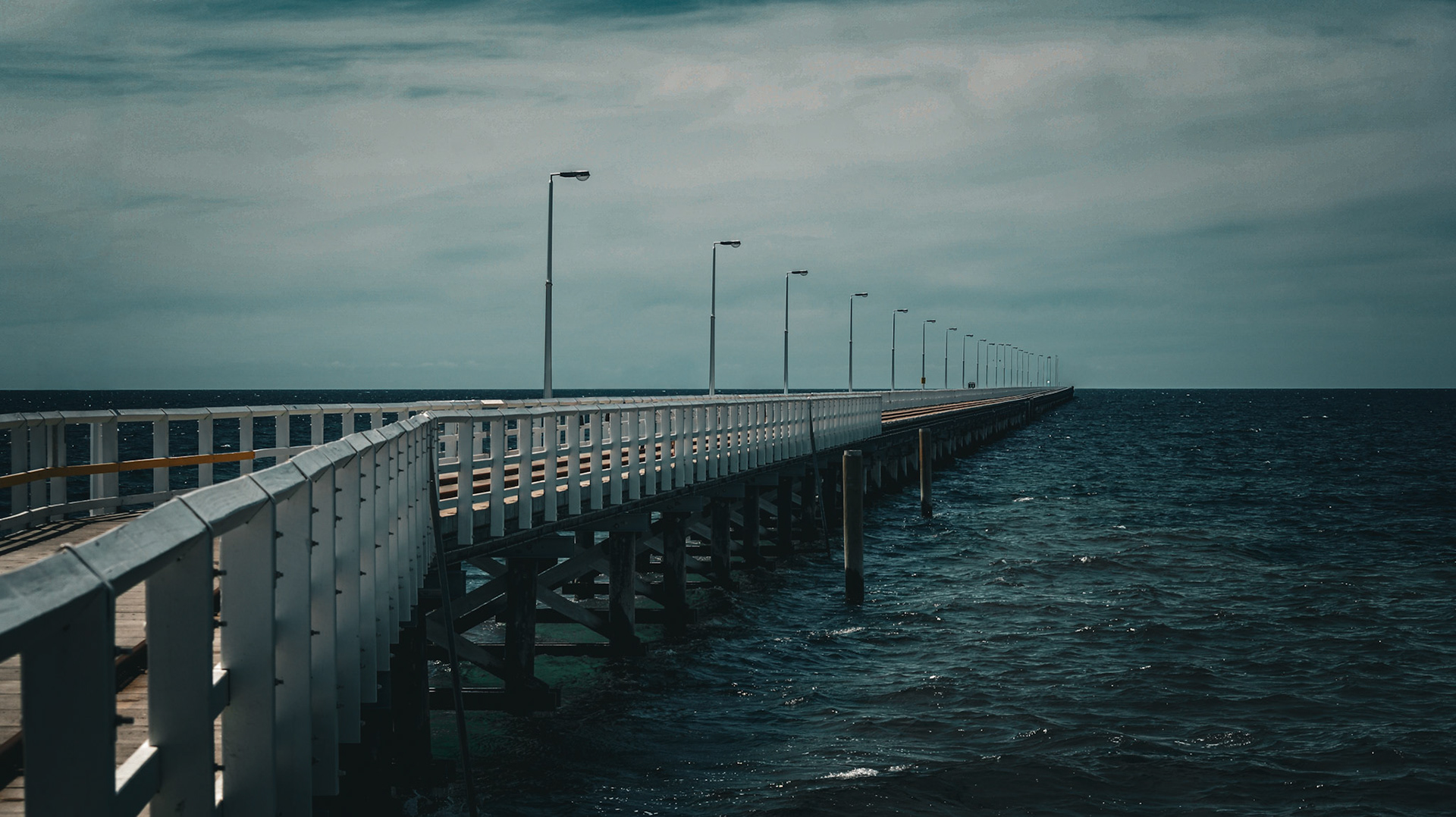 A long wooden pier stretches into the dark ocean, surrounded by a moody sky. Streetlights line the pier, enhancing the serene atmosphere.