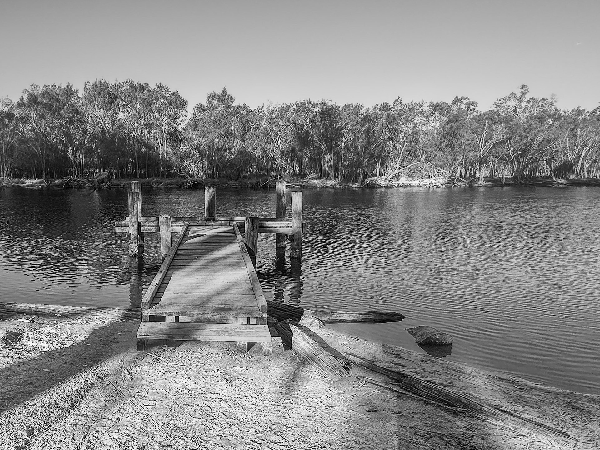 Black-and-white photo of a weathered wooden jetty leading out over calm river water toward a tree-lined opposite bank. Soft ripples reflect the shoreline and nearby posts, while sandy ground, scattered logs and long shadows frame the foreground, creating a peaceful, rustic atmosphere.