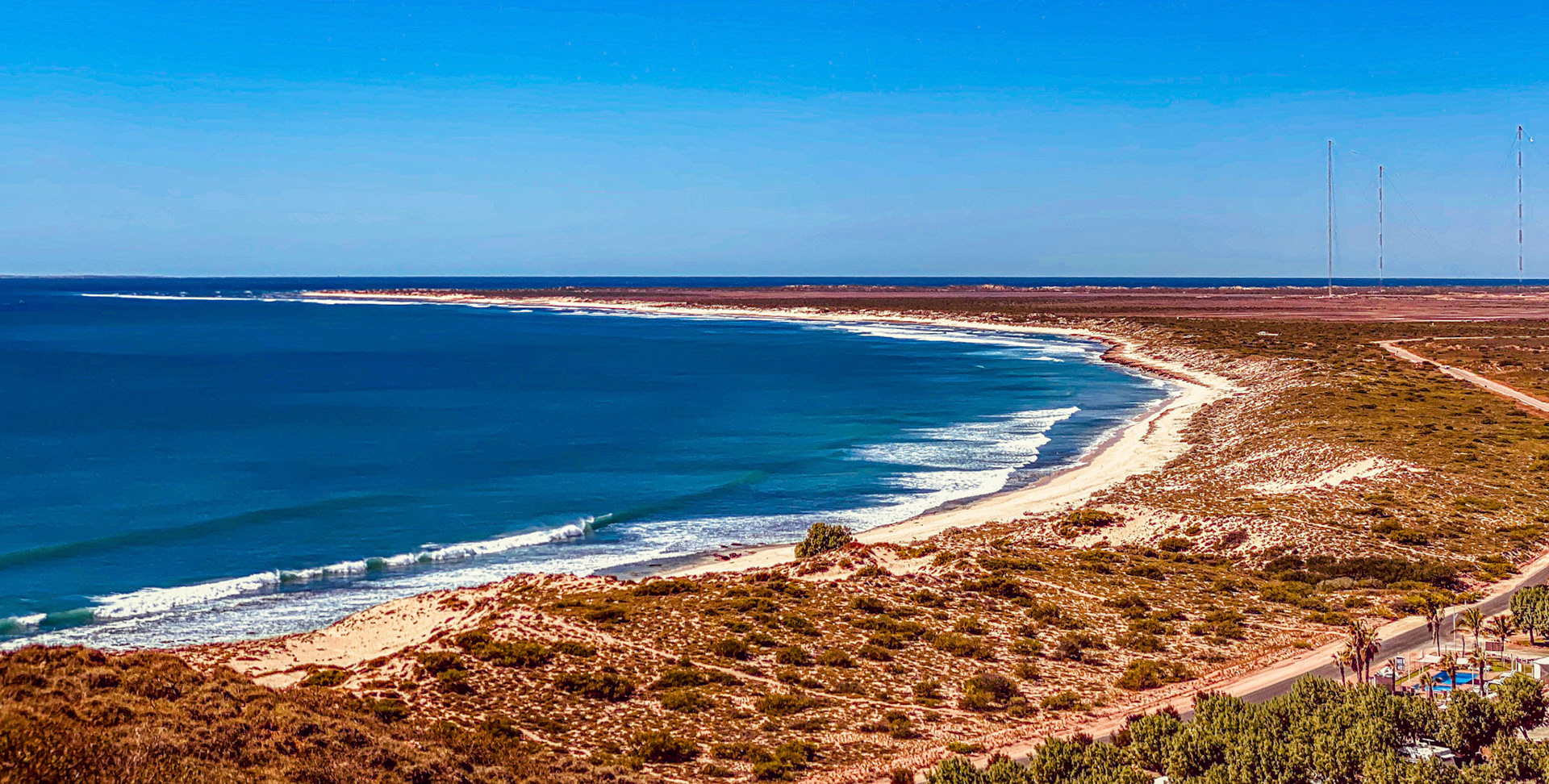 A sweeping view showcases the vast coastline, where waves meet the beach under a clear blue sky, evoking serenity and natural beauty.