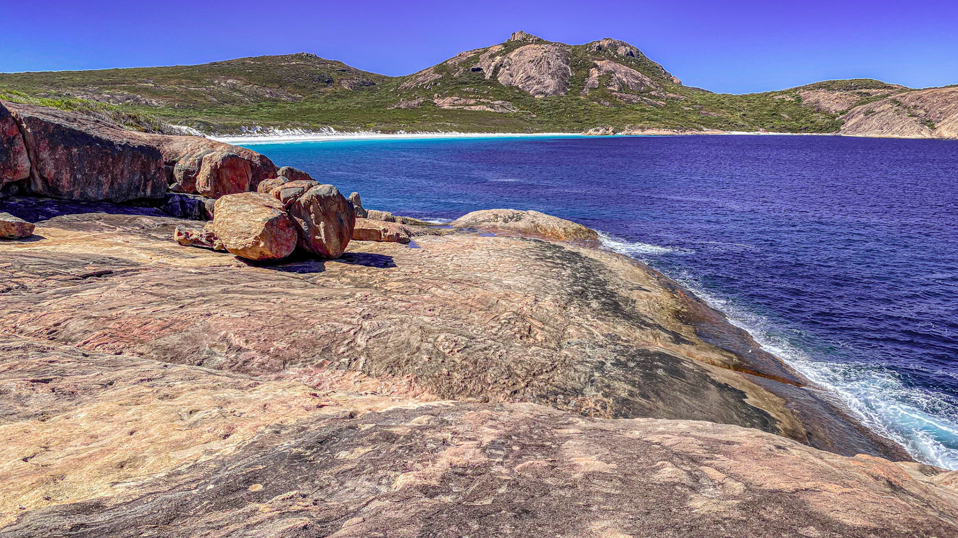 A panoramic coastal view showcases rugged rocks meeting the vibrant ocean, framed by lush mountains and a serene beach.