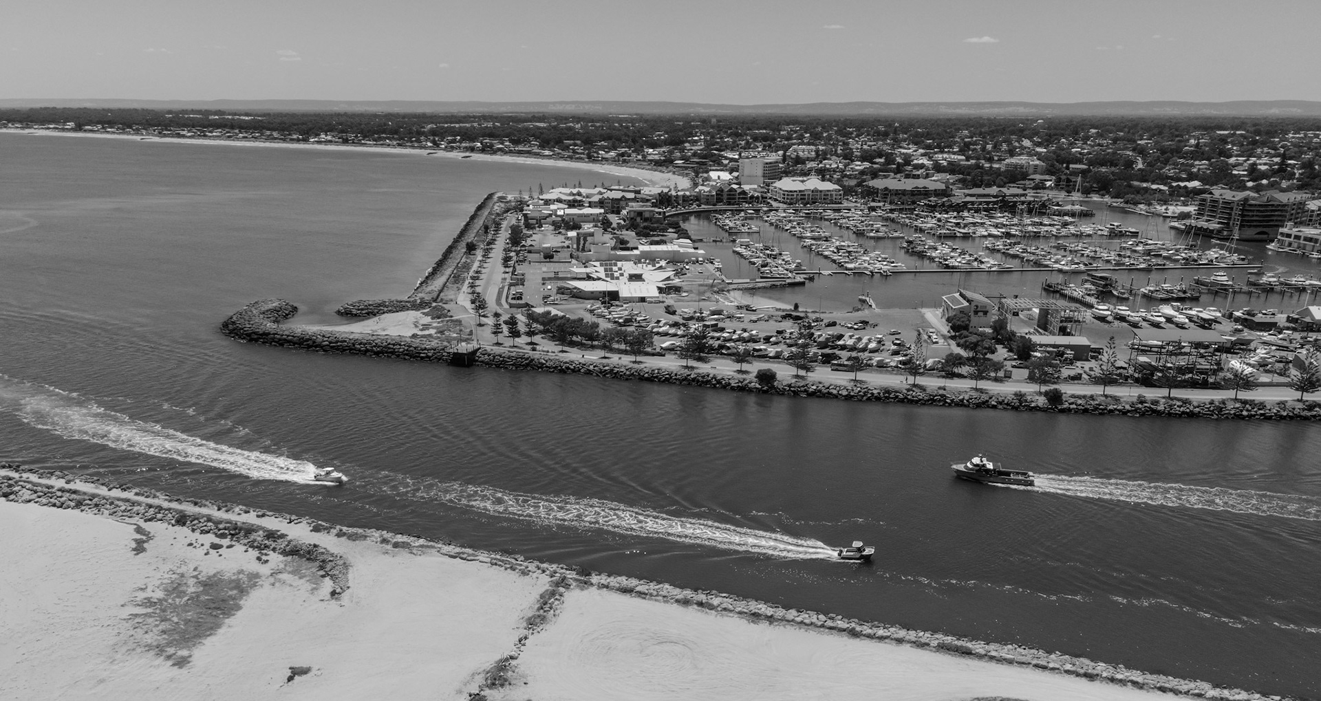 A sweeping black-and-white aerial photograph shows a sheltered marina bordered by long rock breakwaters. A narrow channel runs between curved stone walls as three small motorboats speed through, leaving crisp V-shaped wakes. Inside the harbor, dense rows of berths hold yachts and powerboats, with waterfront buildings, car parks, and a tree-lined road along the spit. Beyond, a low-rise coastal town and sandy beaches stretch to the horizon under a clear sky.