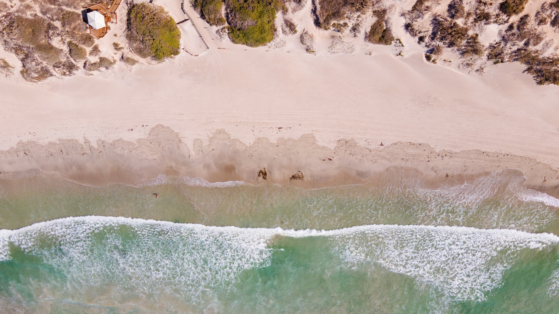 An aerial perspective showcases a serene beach with gentle waves, sand dunes, and tranquil waters, embodying nature's beauty and peace.