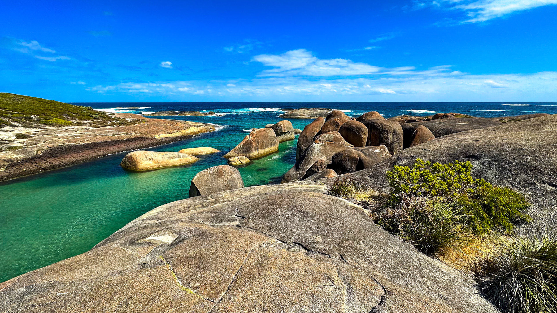 A panoramic view showcases a coastal landscape with turquoise waters, rugged rocks, and a clear blue sky, highlighting nature's beauty.
