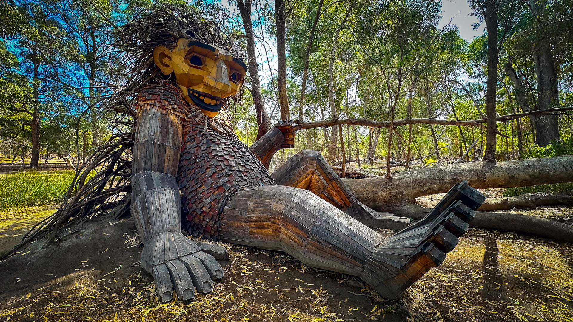 A large wooden sculpture of a friendly troll sits in a sun-dappled forest clearing, showcasing creativity and whimsy.