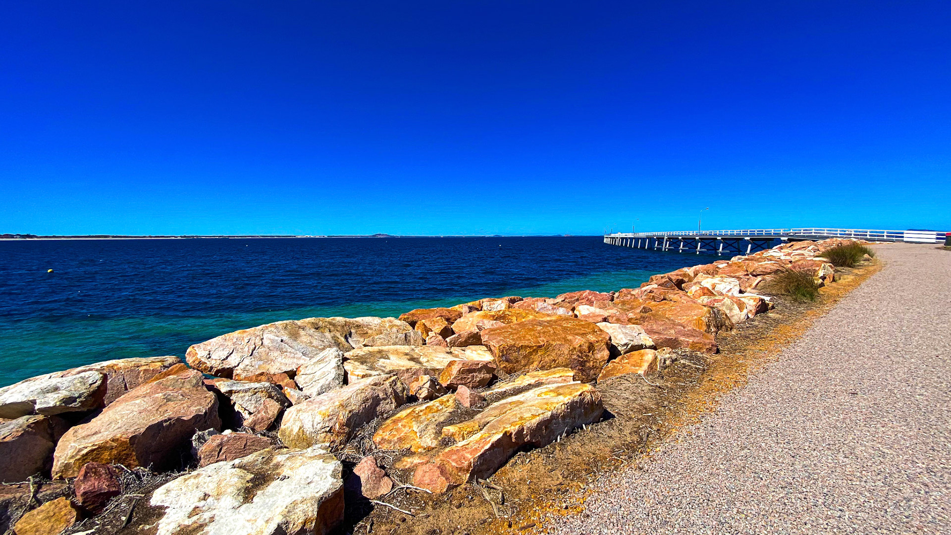A panoramic view showcases a vibrant coastal scene with ocean, pier, and rocks. The clear blue sky enhances the serene atmosphere.
