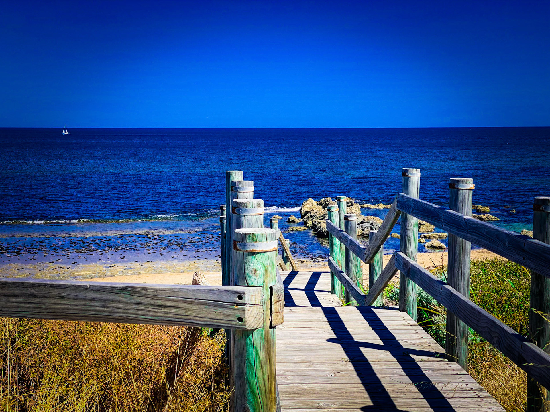 A weathered boardwalk leads to a sandy beach and deep blue ocean under a clear sky, evoking tranquility and escape.