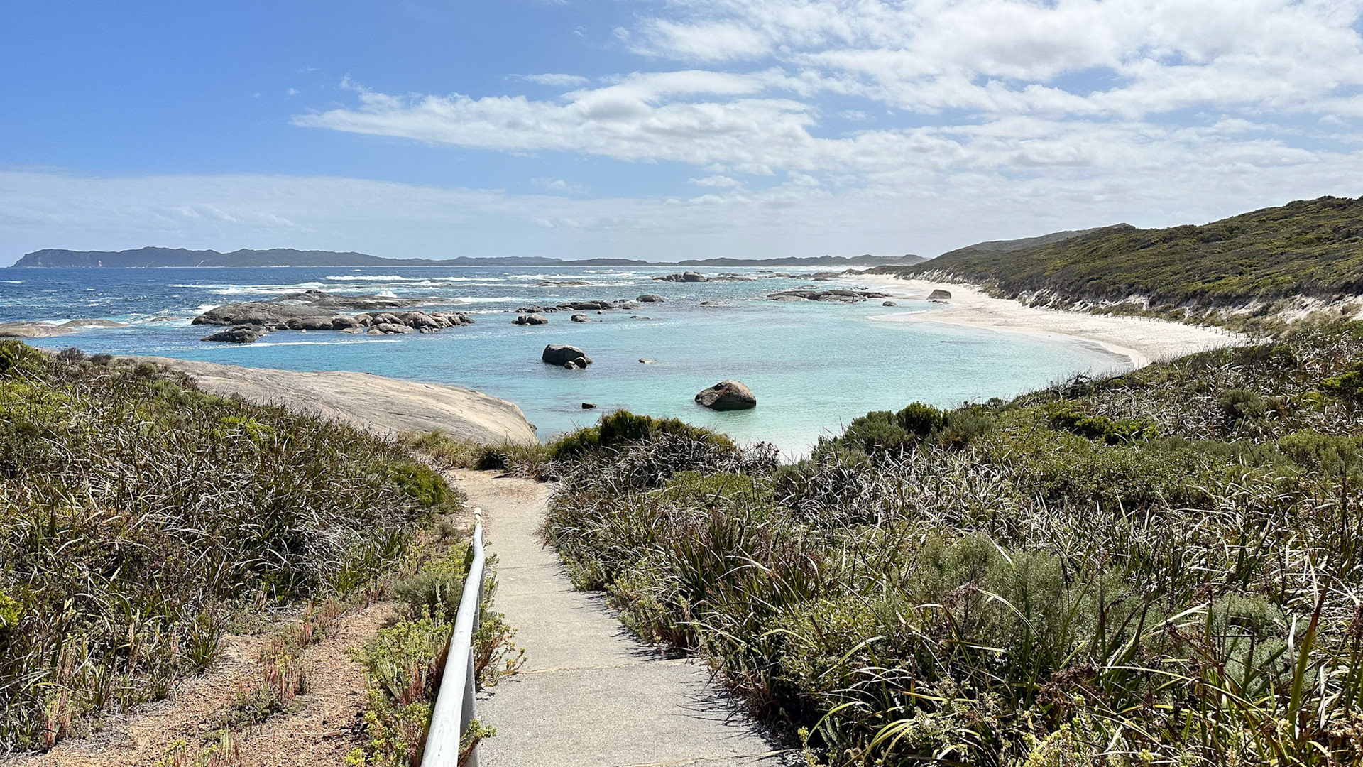 A scenic coastal path descends to a secluded beach, showcasing clear waters and rugged rocks, embodying tranquility and natural beauty.