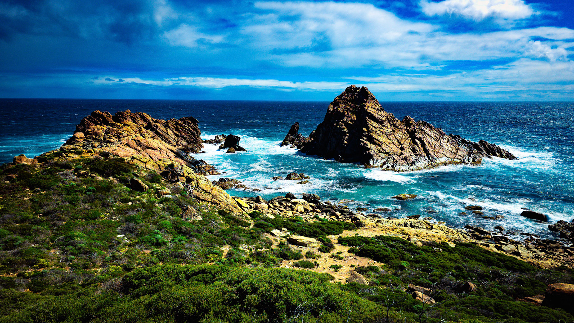 Stunning view of a rugged coastal landscape featuring dramatic rock formations surrounded by the vibrant blues of the ocean. The foreground displays lush green vegetation, contrasting with the deep blue sky and scattered clouds. The image captures the natural beauty and dynamic energy of the sea meeting the land, showcasing the untouched wilderness of coastal regions.