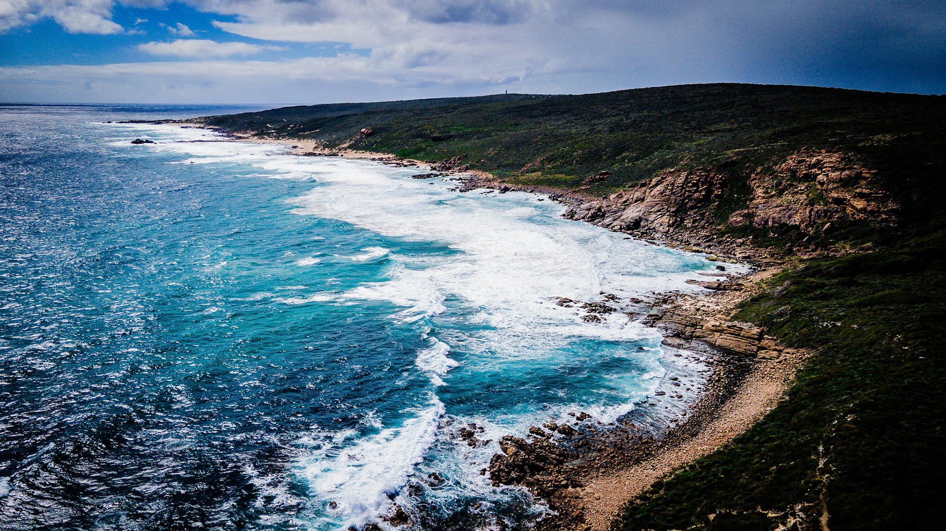 A breathtaking aerial view of a rocky coastline featuring vibrant turquoise waters crashing against rugged cliffs. The scene captures the natural beauty of the ocean meeting the land, framed by a dramatic sky.