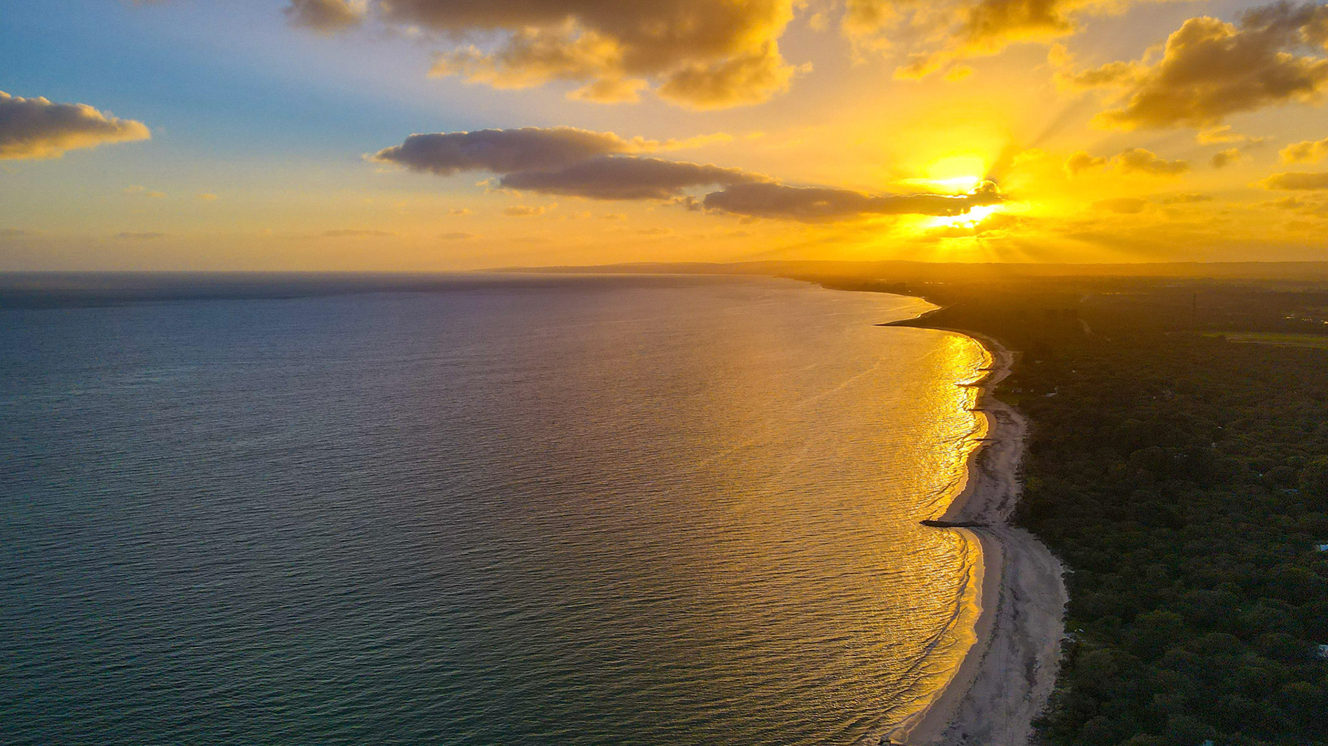 A breathtaking aerial view of a sun setting over the ocean, casting a golden glow across the water and shoreline. The vibrant sky is filled with colorful clouds, creating a serene and tranquil atmosphere. Perfect for themes of nature, travel, and relaxation.