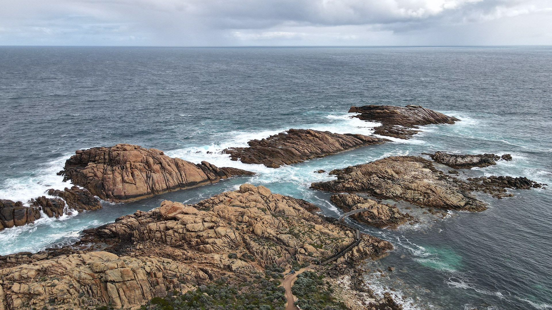 A striking aerial view of a rugged coastal landscape featuring prominent rocky outcrops surrounded by the ocean. The waves crash against the rocks, creating a dramatic seascape. The cloudy sky adds a moody atmosphere to the scene, emphasizing the beauty and power of nature. A narrow path and boardwalk traverse the rocky terrain, offering a sense of scale and human interaction with the natural environment.