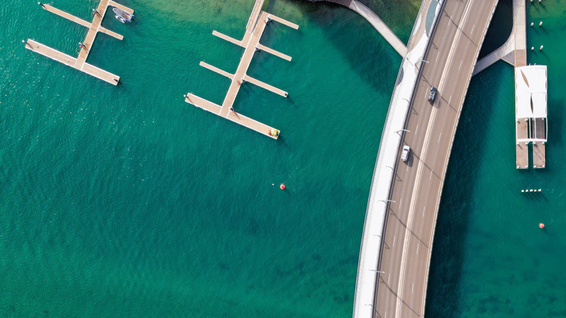Captivating aerial shot displaying a vibrant marina with several boat docks alongside a serene, turquoise body of water. The image also features a smooth highway curving over the marina, showcasing a blend of natural beauty and urban infrastructure. This visually stunning composition is ideal for showcasing waterfront development, recreational boating, and travel destinations. Perfect for real estate marketing, travel blogs, and environmental documentaries.