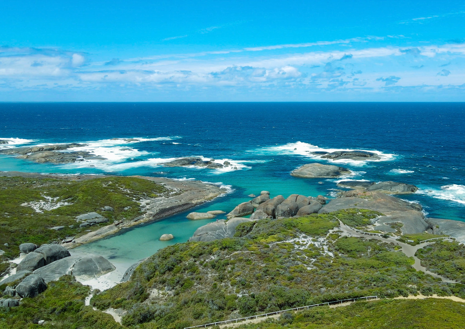 An aerial view showcases a stunning coastal landscape with ocean waves, rocks, and lush vegetation under a clear blue sky.