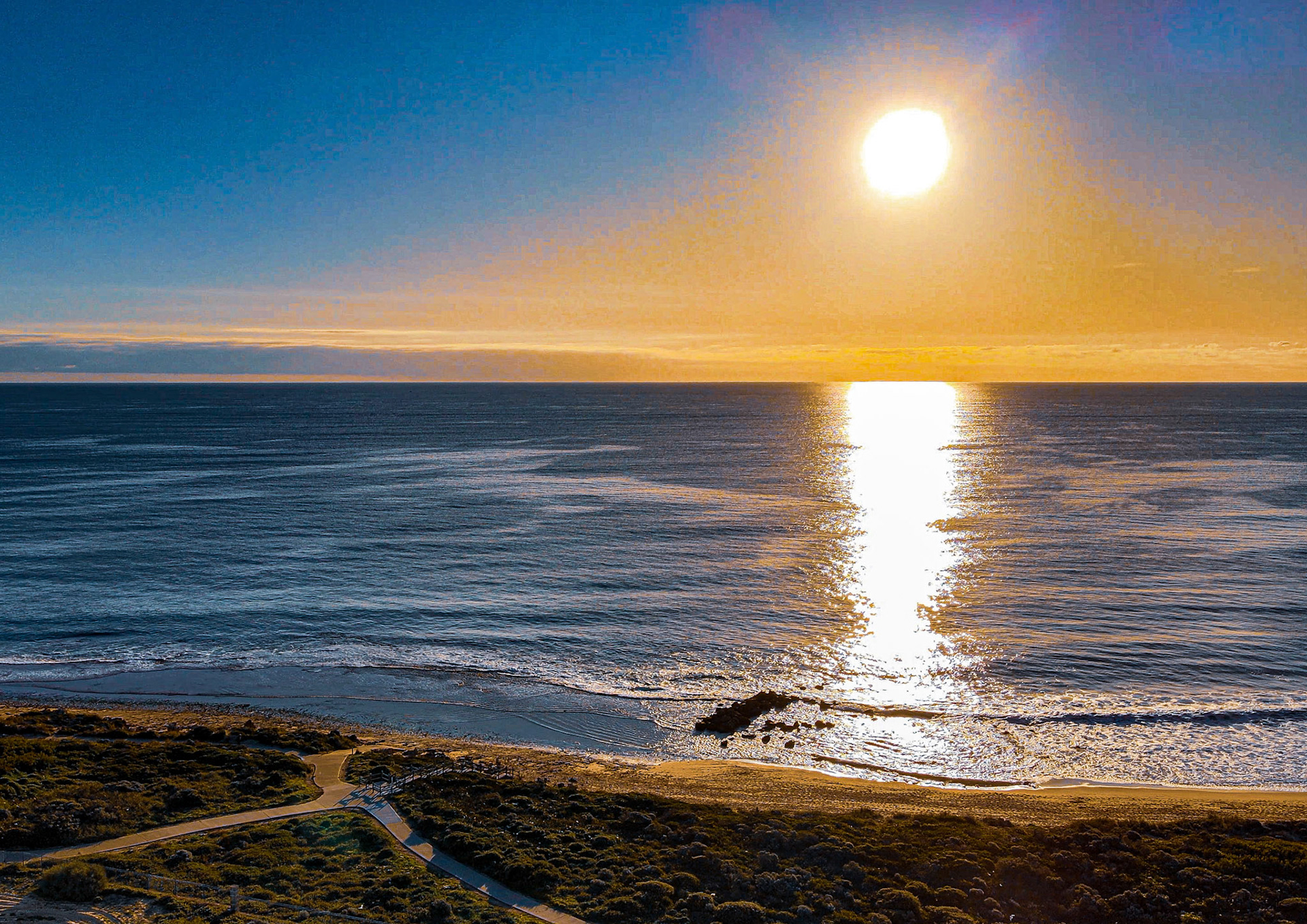 A serene sunset casts vibrant hues over the ocean, reflecting on waves. The peaceful beach path leads through nature's tranquility.