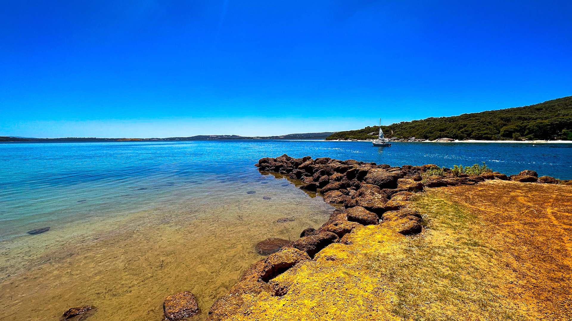 A tranquil coastal landscape features a calm sea, rocky shore, and a sailboat under a clear blue sky, evoking serenity and peace.