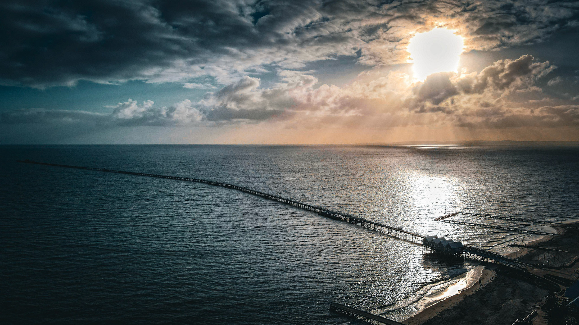 Captured in stunning detail, this aerial shot showcases a long pier extending into the ocean under dramatic skies. The sun breaks through heavy clouds, casting brilliant rays and reflections over the water's surface, creating an awe-inspiring natural scene.