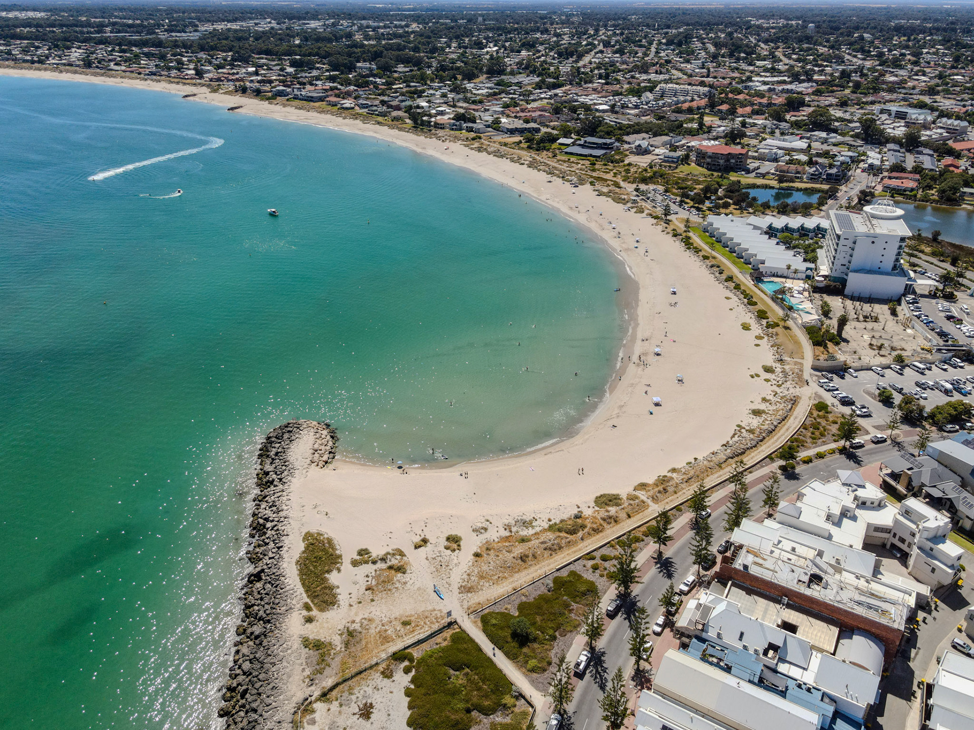 A high-angle aerial photo of a crescent-shaped sandy beach with clear turquoise water. A rock breakwater juts into the bay, forming a sheltered cove where a few small boats and swimmers are visible. A jet ski leaves a curved white wake across the water. Scattered beachgoers, umbrellas and towels dot the shoreline. Behind the beach are a promenade, parking areas, a cluster of low-rise buildings and a multi-story hotel, with residential streets and greenery extending inland under bright daylight.