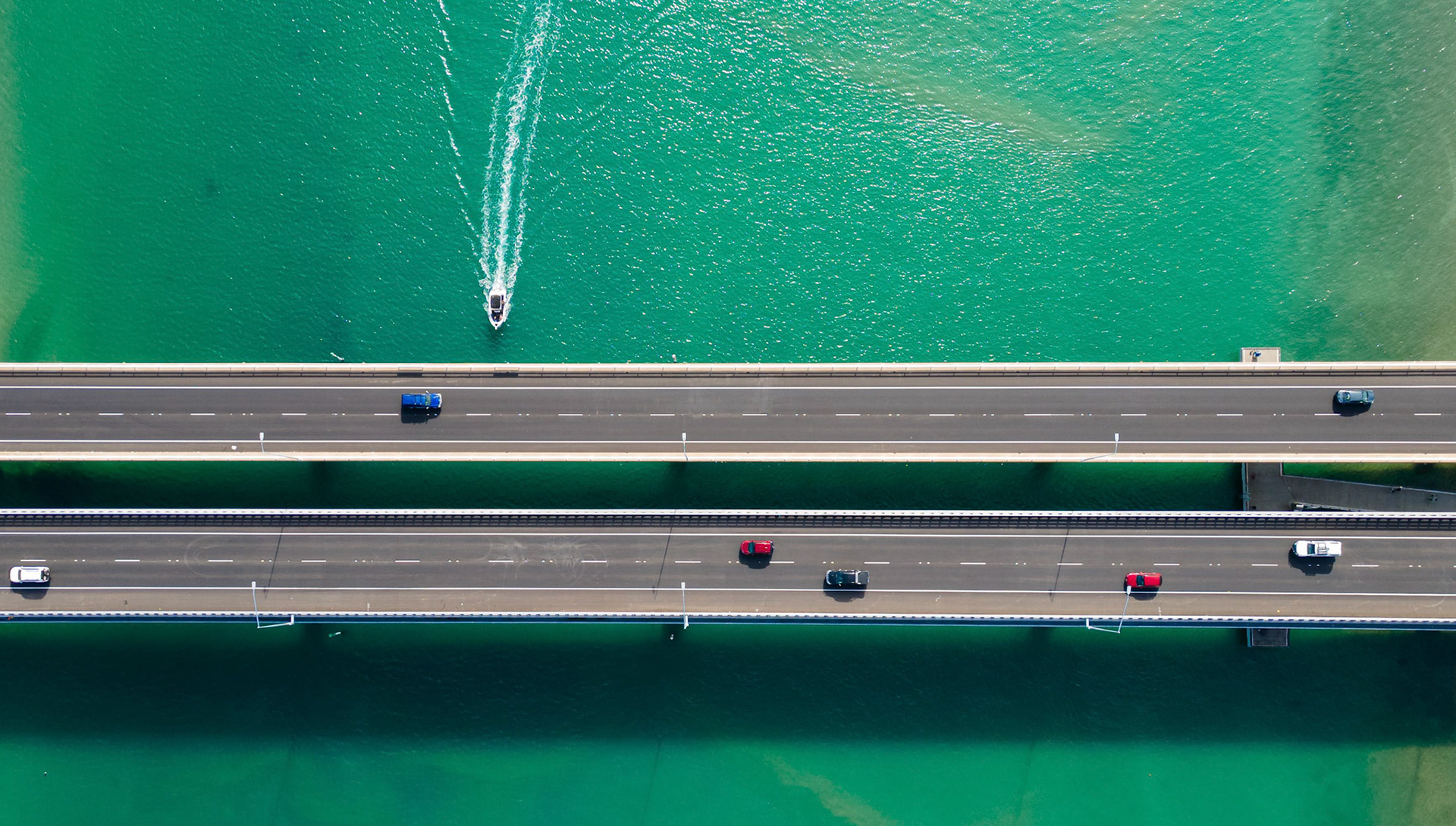 Stunning aerial view capturing two parallel bridges spanning across vibrant turquoise waters. The image showcases a dynamic scene with vehicles traversing the roadways while a boat glides smoothly along the water below. Ideal for travel, infrastructure, and environmental themes, this captivating composition highlights the intersection of nature and modern engineering, emphasizing the beauty of natural landscapes and human construction.