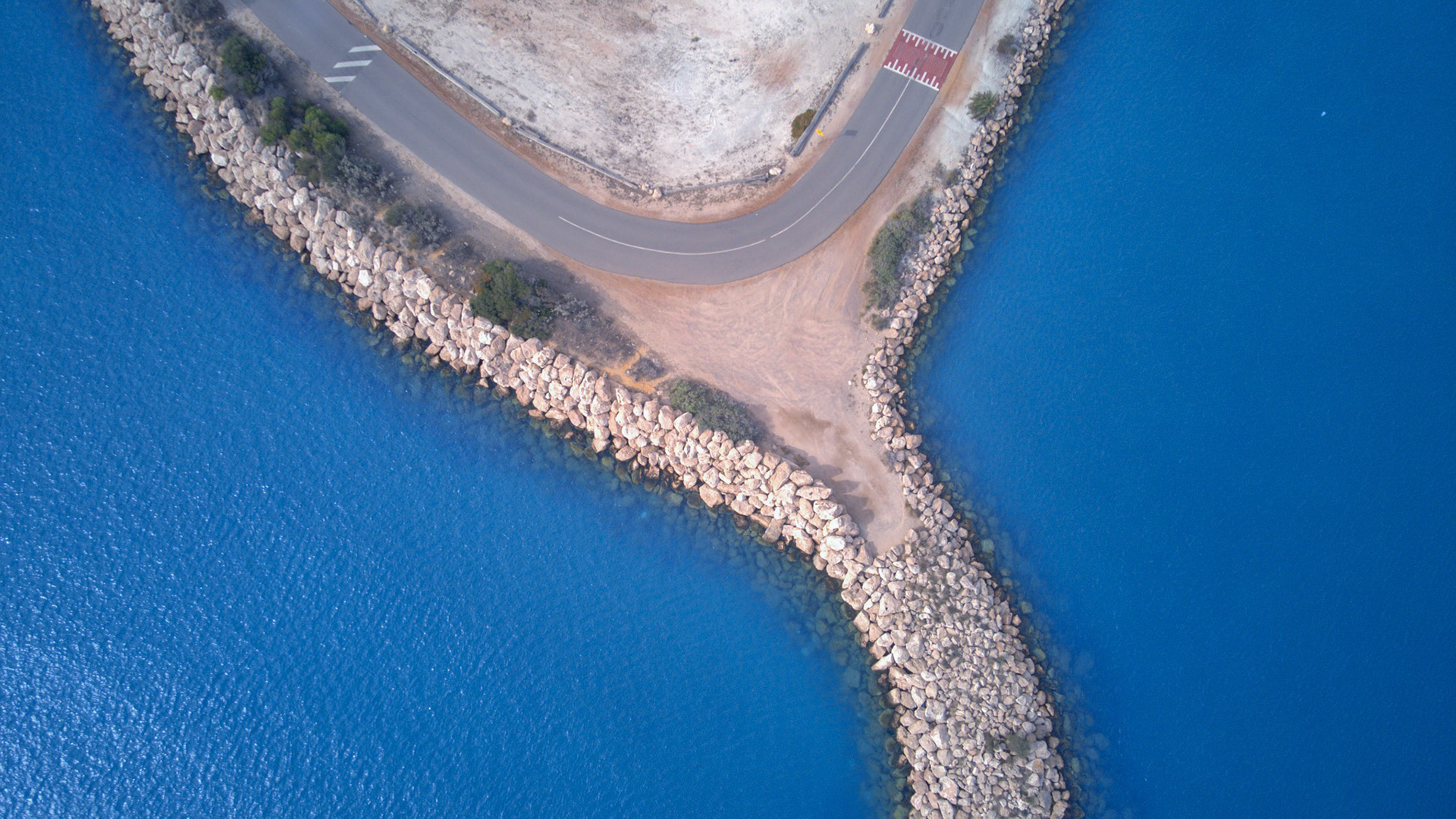 An aerial view showcases a curving road along a rocky breakwater, dividing the deep blue sea and highlighting natural beauty.