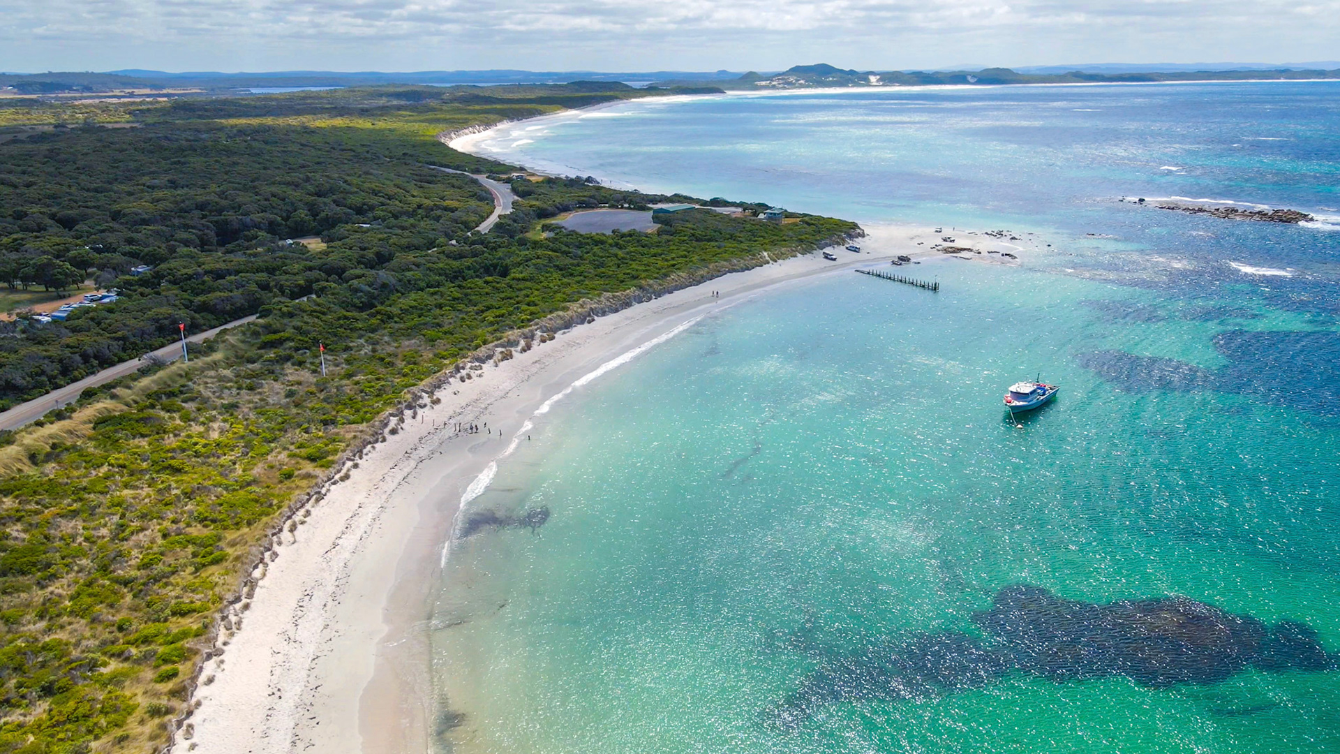 This aerial view showcases a tranquil beach meeting the calm ocean. Lush trees and a small boat enhance the serene coastal landscape.