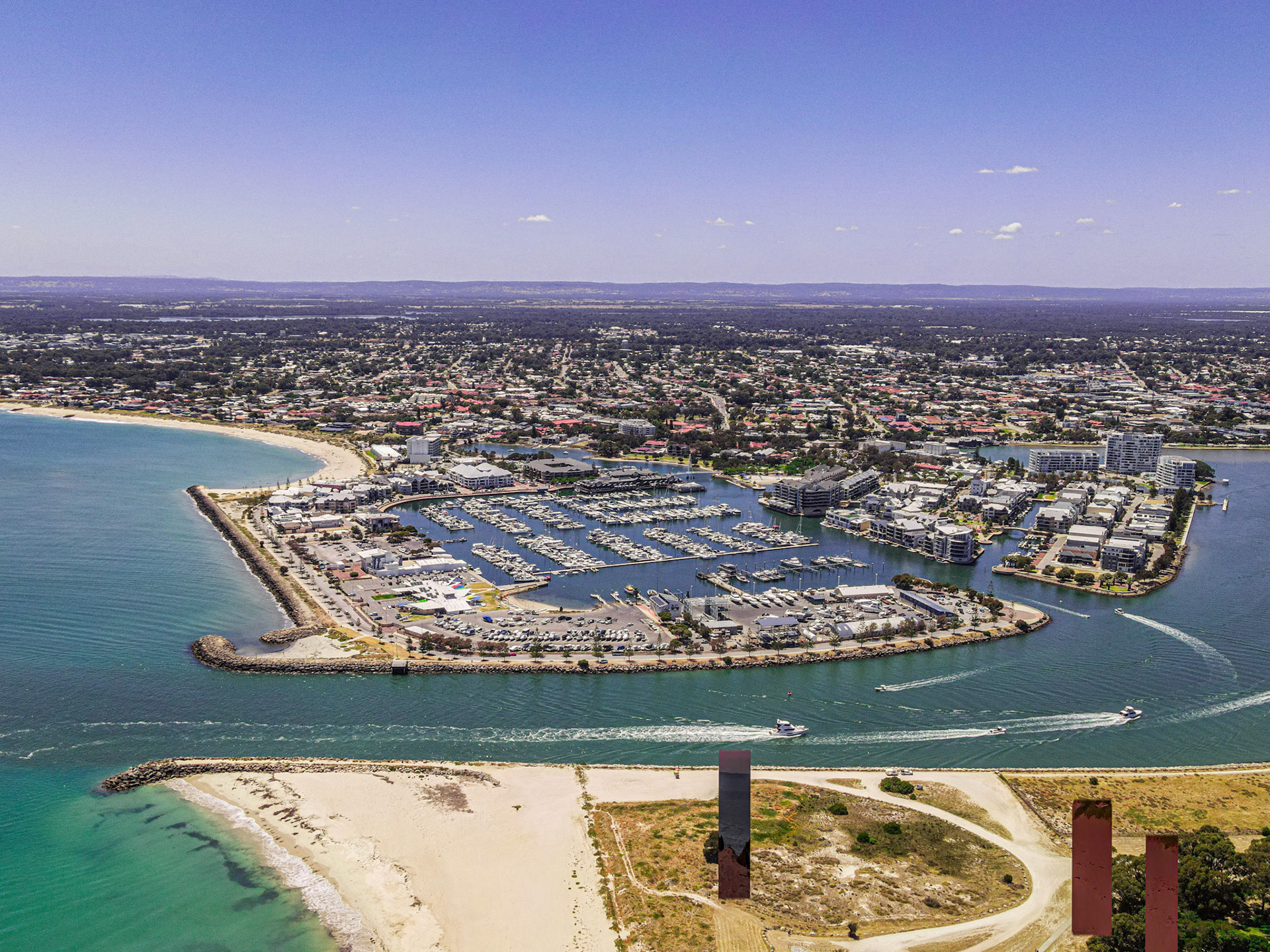 An elevated view of a sunny coastal city featuring a sheltered marina and winding canals lined with apartments and waterfront homes. White-sand beaches and rock breakwaters frame turquoise water, while several small boats carve wakes through the main channel. The urban area stretches inland beneath a clear blue sky.