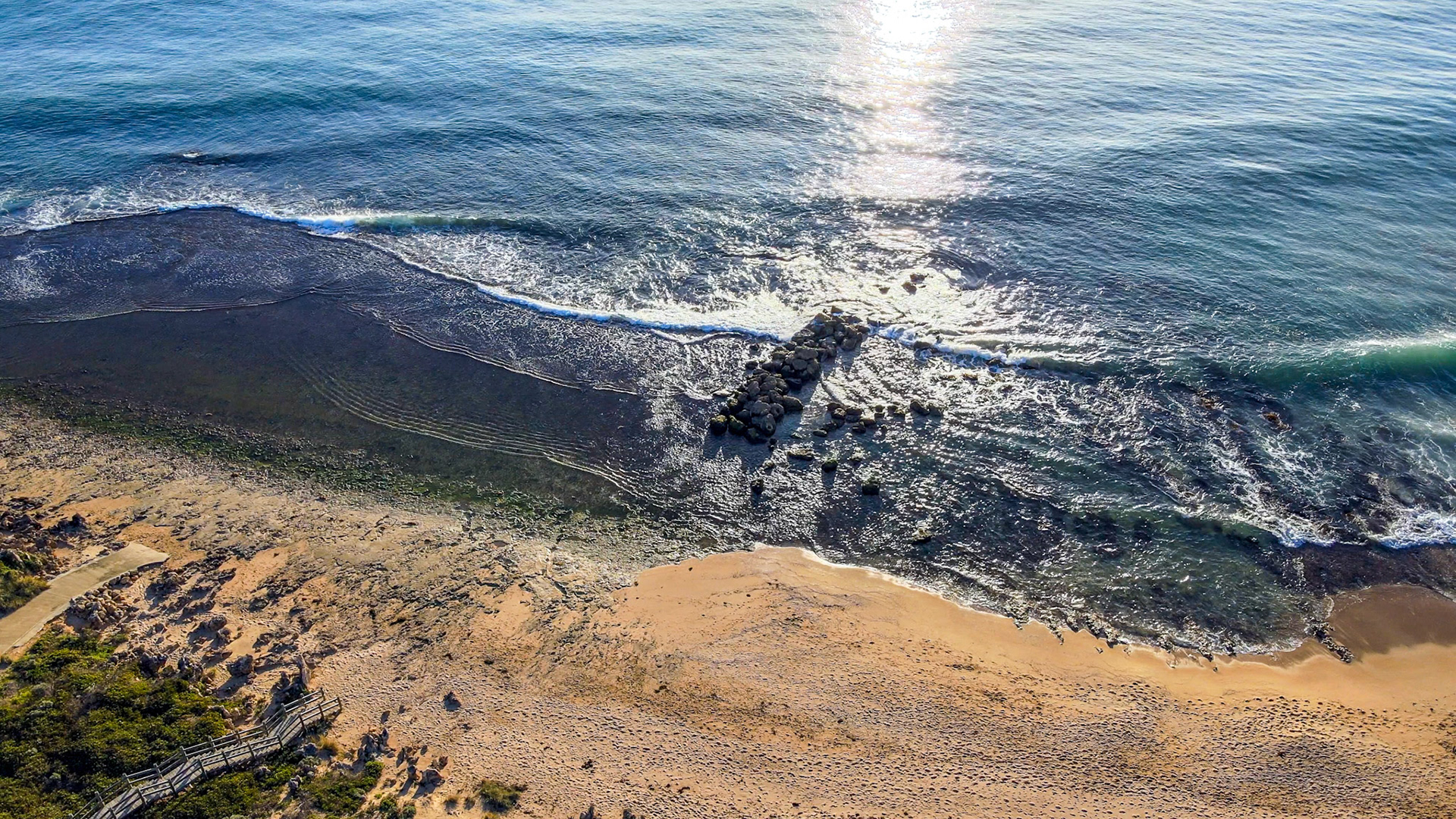 An aerial view showcases the serene meeting of ocean and coastline, with gentle waves lapping against rocky formations and sandy shores.