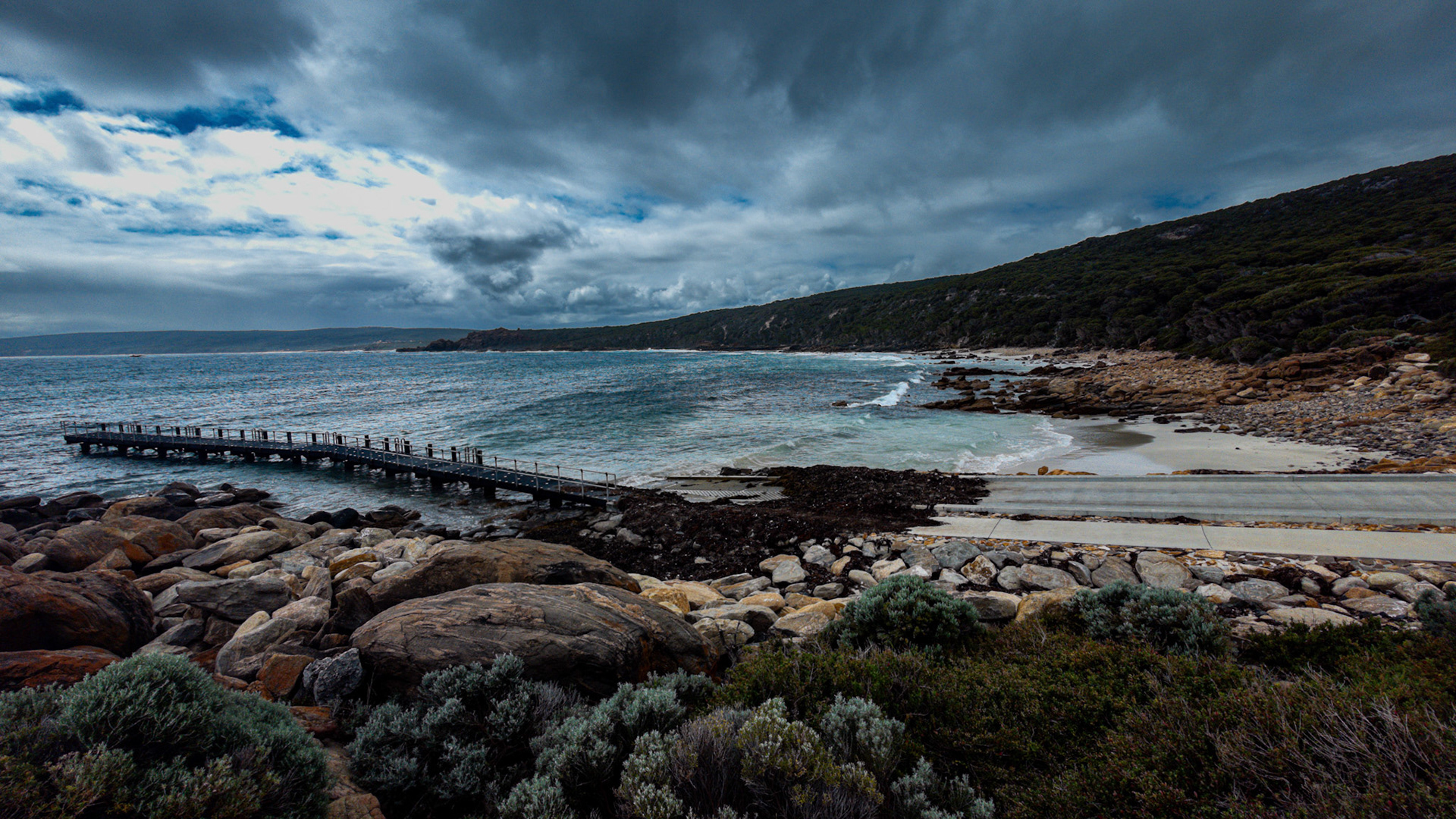 A breathtaking view of a rugged coastal landscape featuring a wooden pier extending into the ocean under a dramatic, cloudy sky. The rocky shoreline and lush greenery create a natural contrast with the deep blue sea and white surf. This serene image captures the untamed beauty of nature and is ideal for use in travel, adventure, and nature-focused designs.