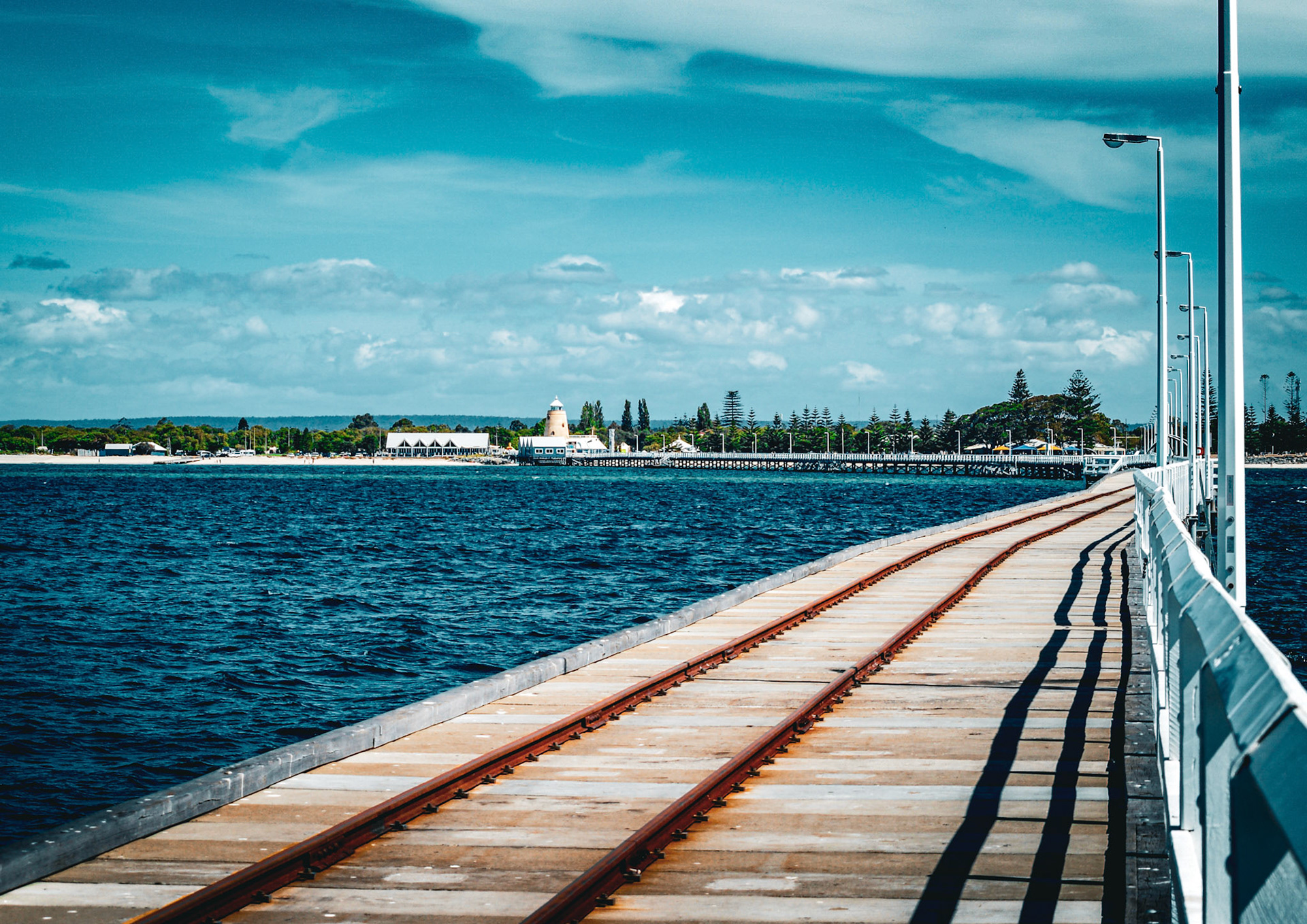 A wooden pier with rusty train tracks leads over the ocean, showcasing a serene coastal landscape and distant shore with a lighthouse.