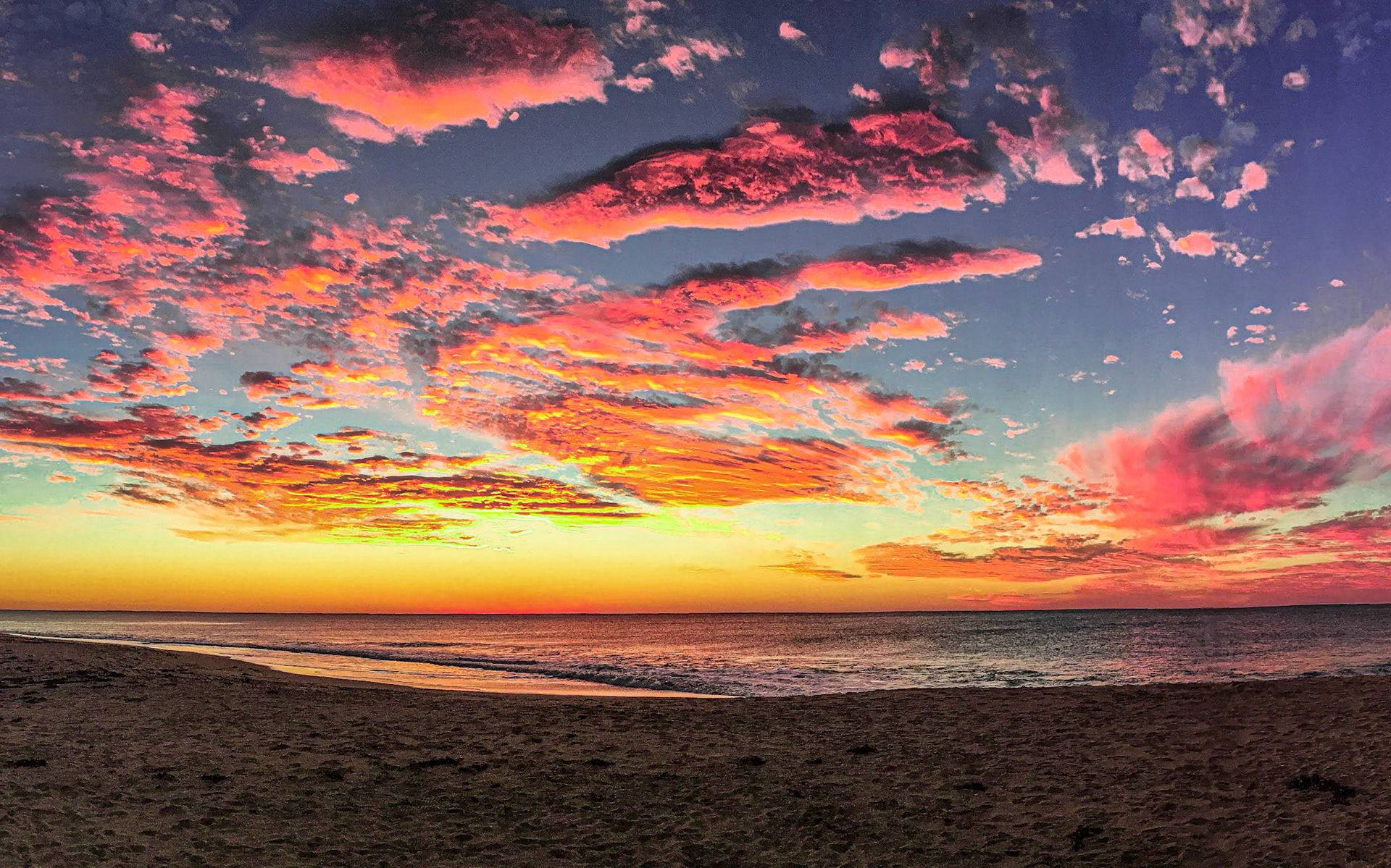 A panoramic view showcases a stunning sunset over the ocean, with vibrant colors reflecting on the calm sea and sandy beach.