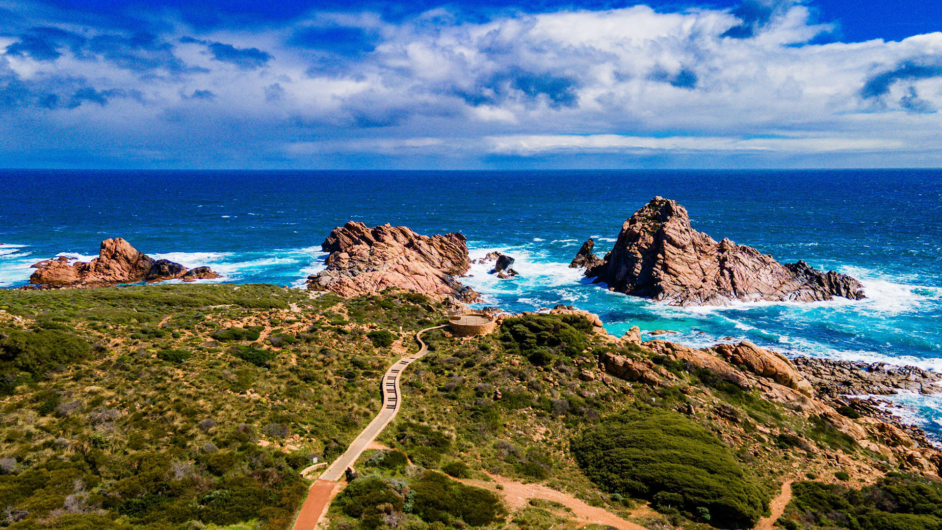 A breathtaking aerial view of a rocky coastal landscape, featuring rugged rock formations surrounded by the vibrant blue ocean. The foreground showcases lush green vegetation and a winding path leading to a viewing platform, adding depth to the natural scenery. The sky is filled with dramatic clouds, enhancing the beauty of this captivating seaside vista.