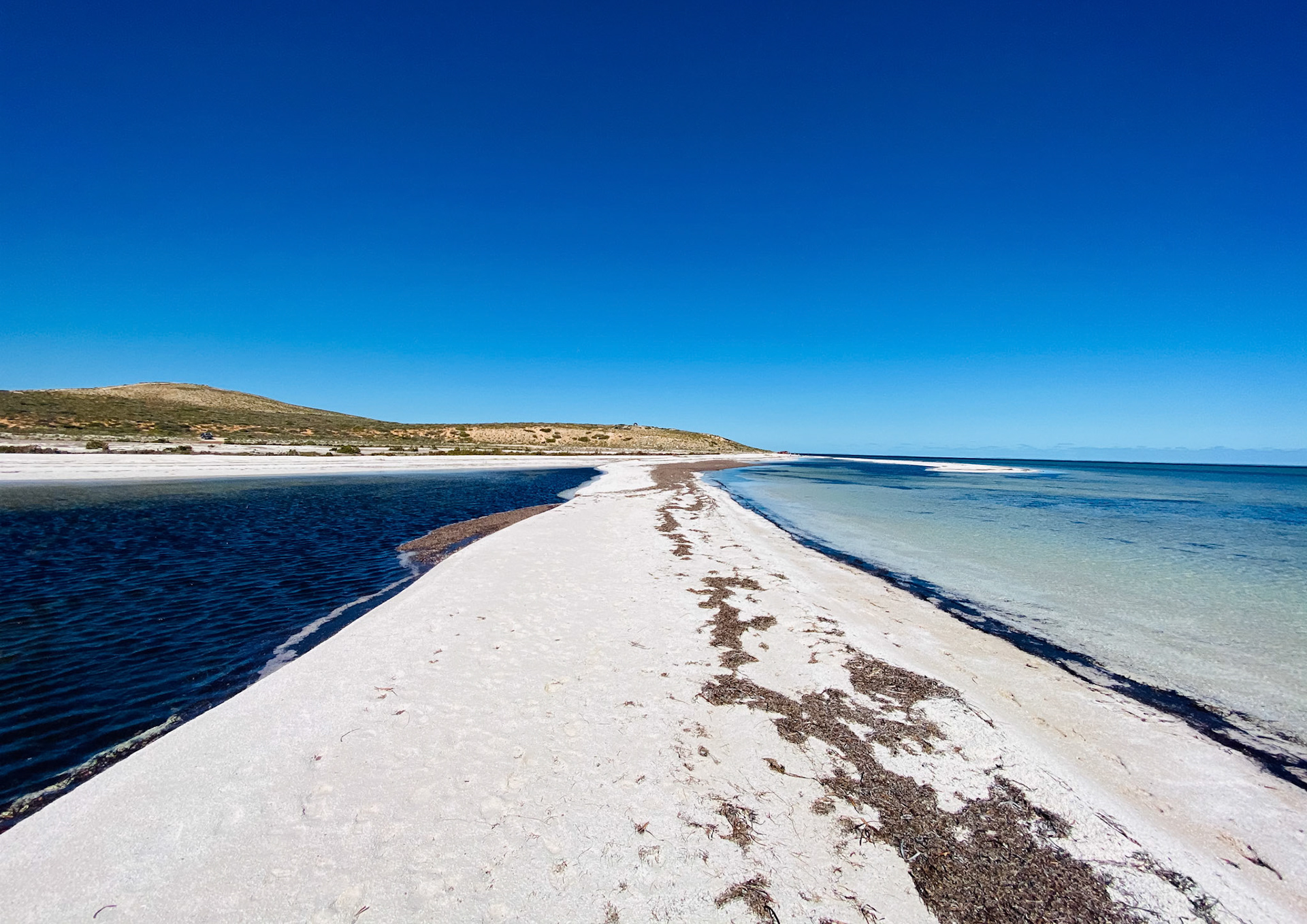 A serene beach landscape features a narrow strip of white sand, dark water, and a clear blue sky, evoking tranquility and natural beauty.