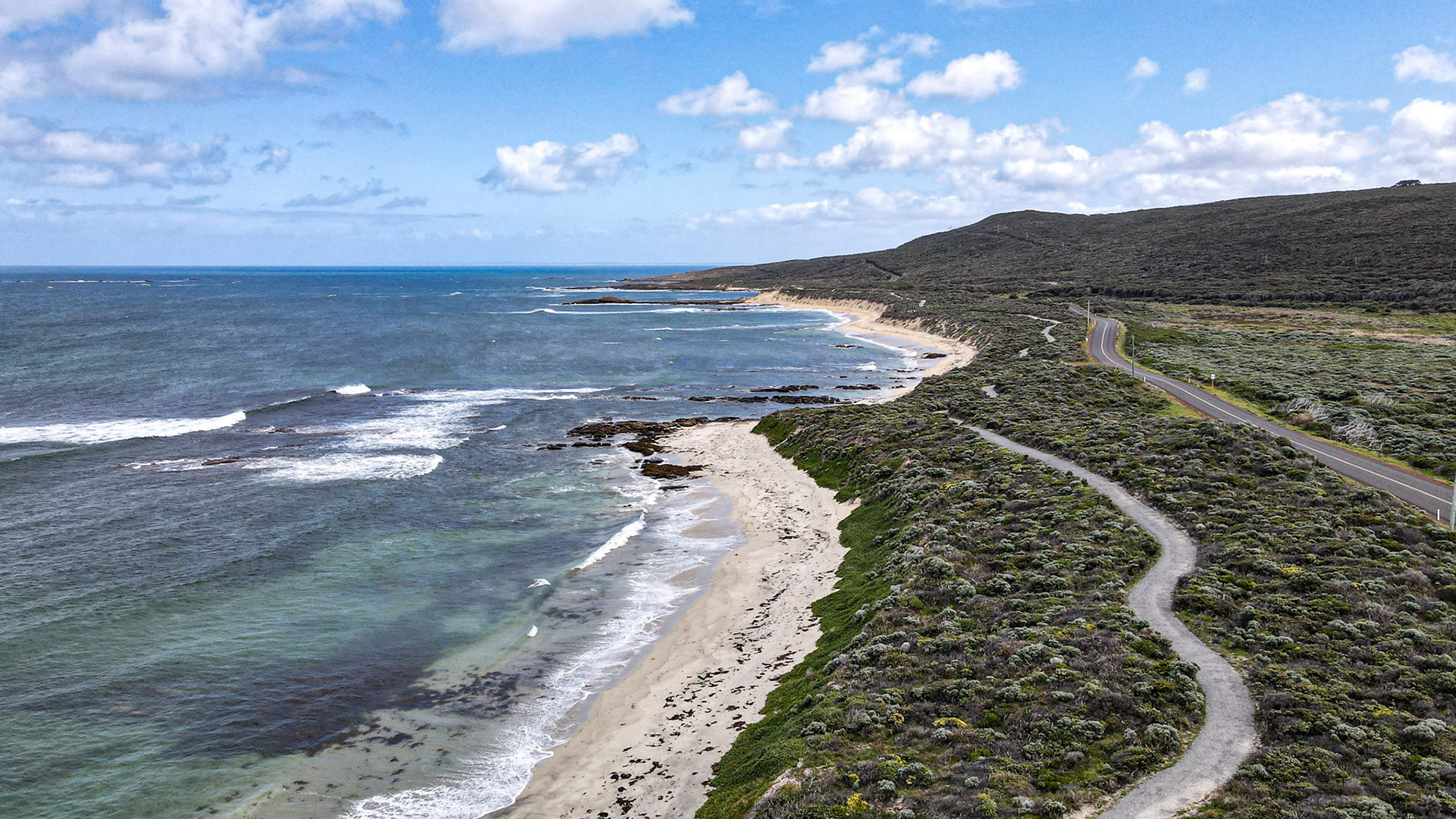 A stunning aerial view of a picturesque coastal road winding along a rugged coastline with lush greenery. The image captures the natural beauty of the seascape, featuring white sandy beaches, rolling waves, and a clear blue sky dotted with fluffy clouds. This serene and tranquil setting is ideal for travel and nature-themed projects, showcasing the untouched charm of a coastal landscape.