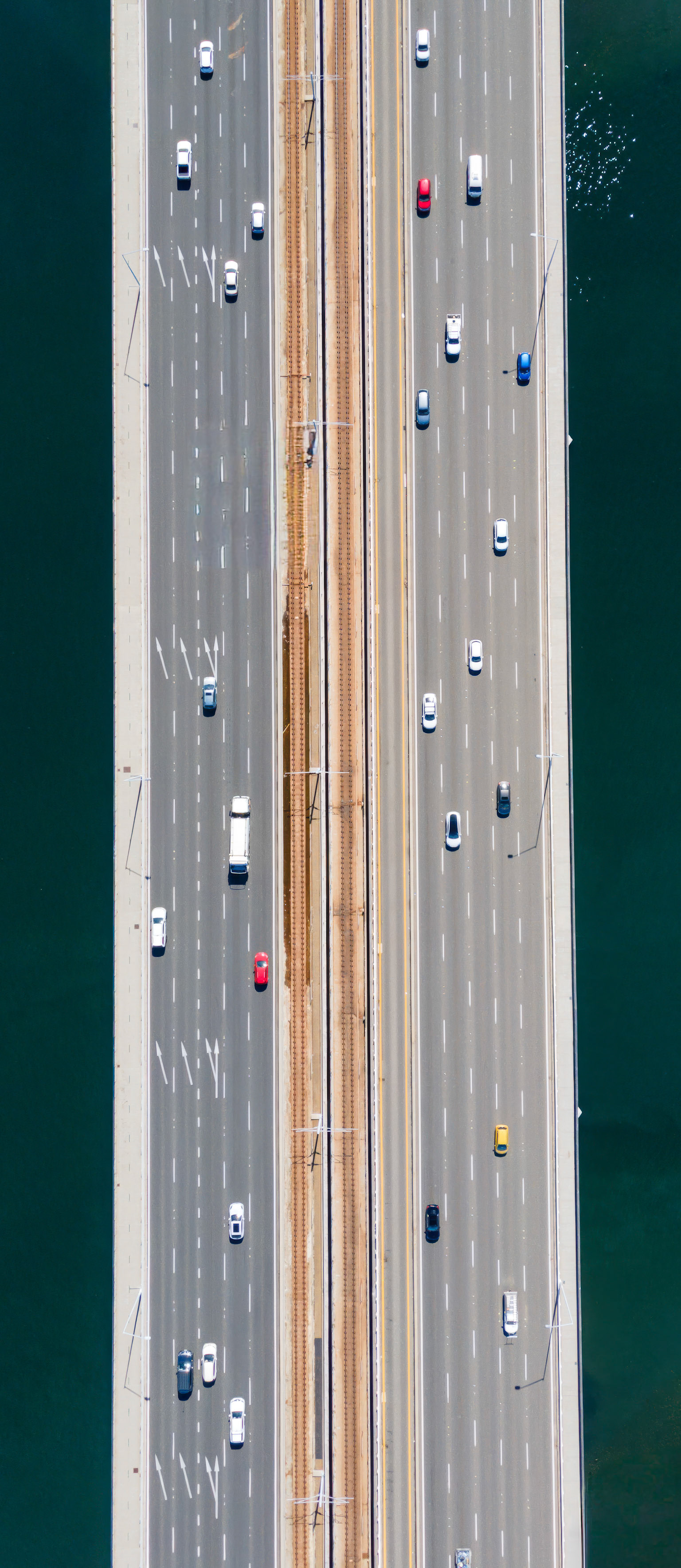 A vibrant scene shows cars traveling on a wide highway alongside a railway track. Below stretches a sparkling body of water, reflecting the bright sky.