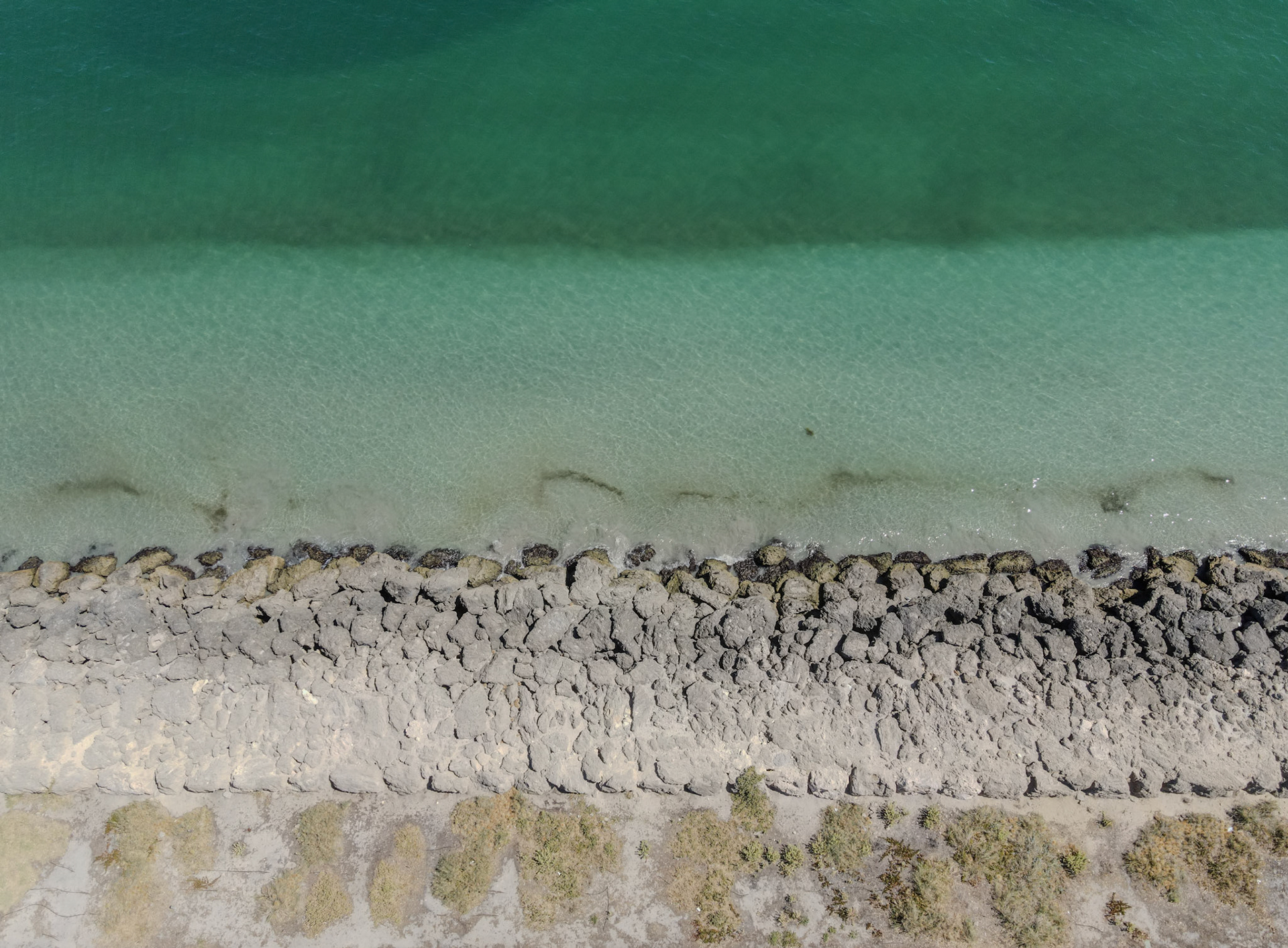 A top-down aerial view captures a rocky seawall bordering clear, shallow water that shifts from pale aqua near the shore to deeper green offshore. The riprap forms a rugged line between the sea and a sparse strip of coastal vegetation, creating clean, parallel bands of texture and color. Gentle ripples and submerged sand patterns are visible through the translucent water.
