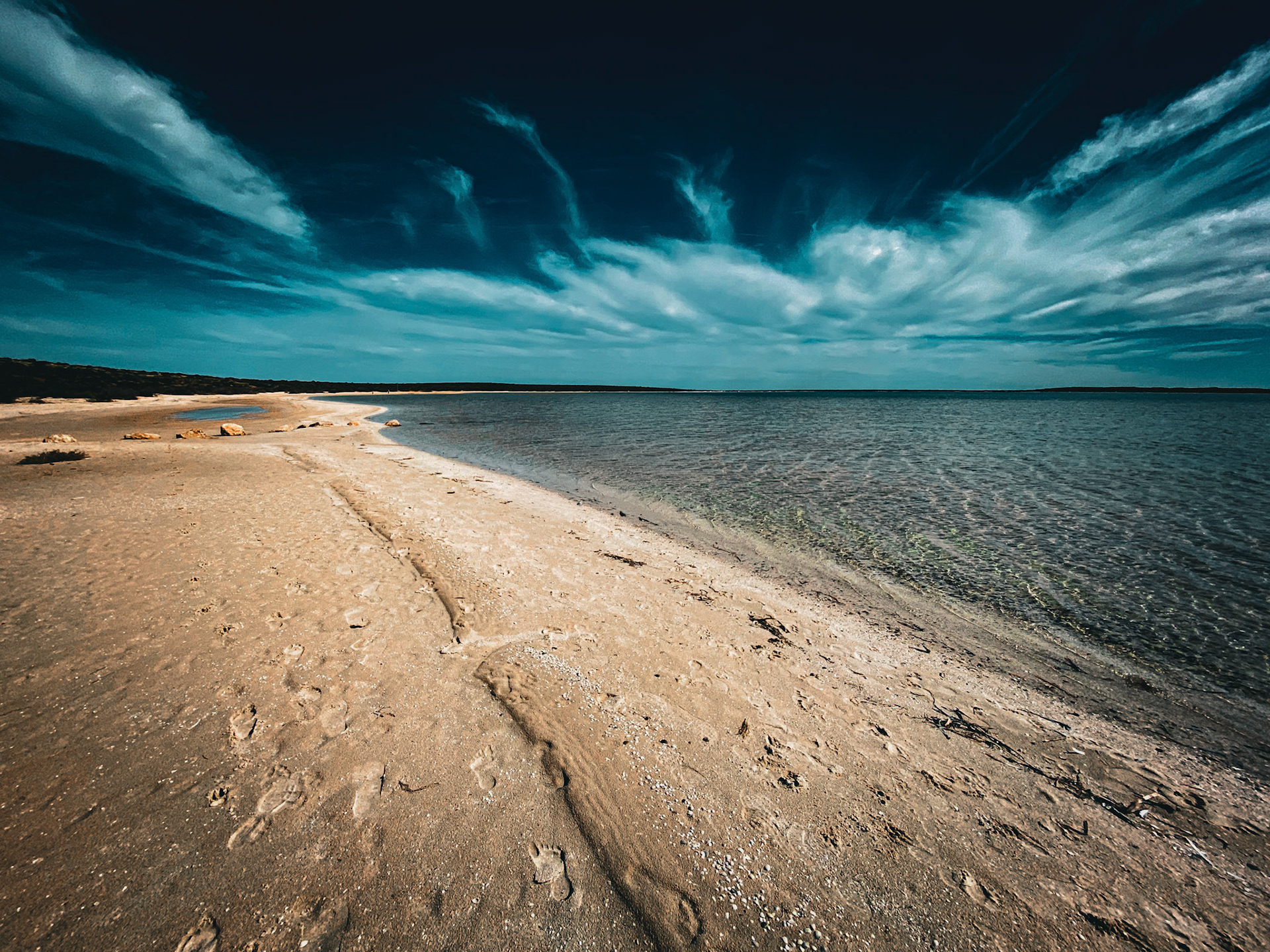 A serene beach landscape showcases calm waters and a cloud-streaked sky. Footprints in the sand hint at recent visitors.