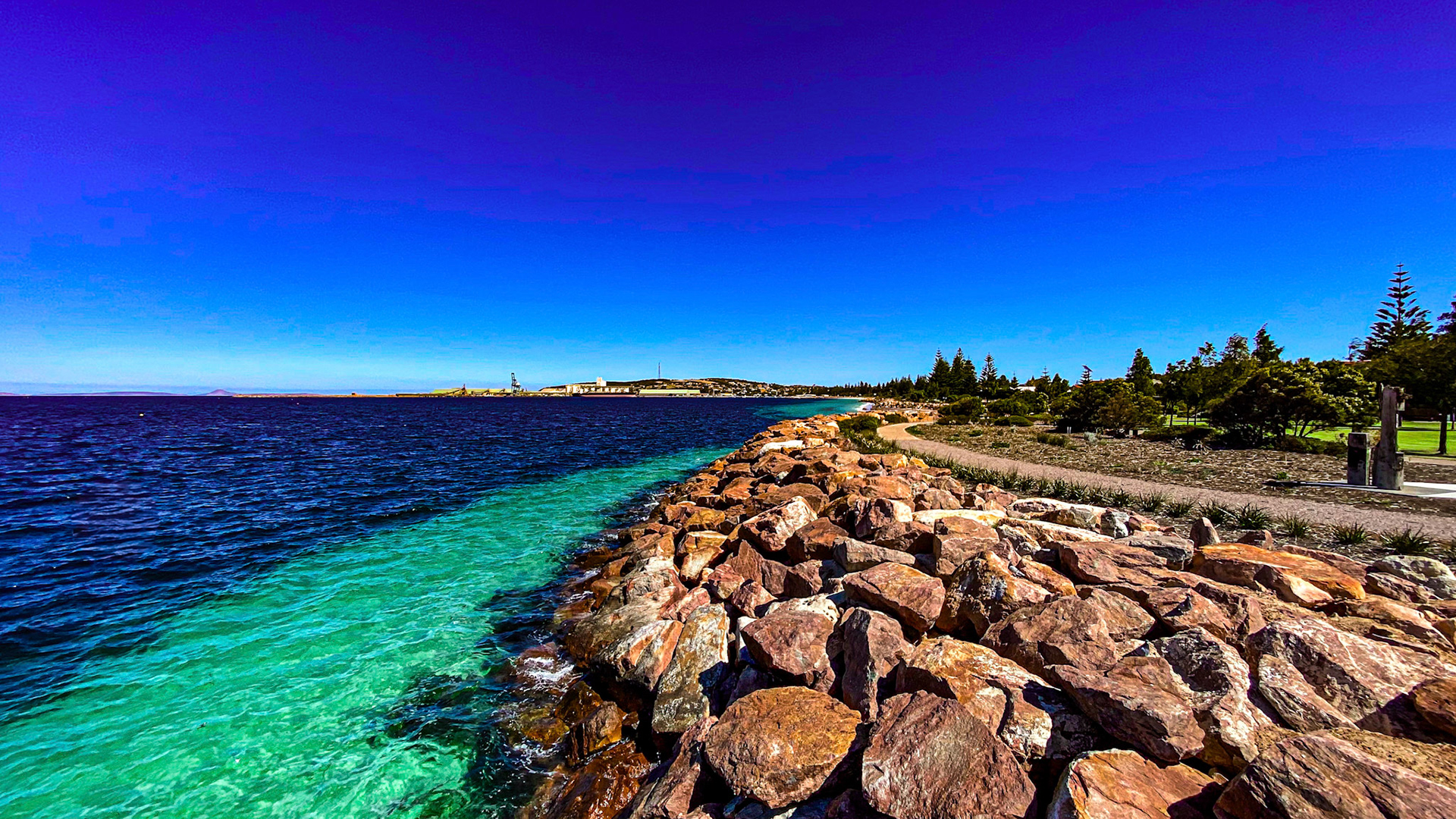 A vibrant coastal scene features deep blue ocean waters, a rocky breakwater, and a tranquil town under a clear blue sky.