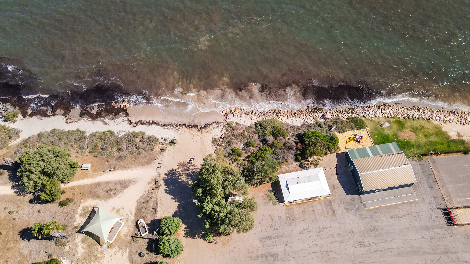 An aerial view showcases a coastal landscape where the beach meets the ocean. Buildings and a canopy are visible near the shoreline.
