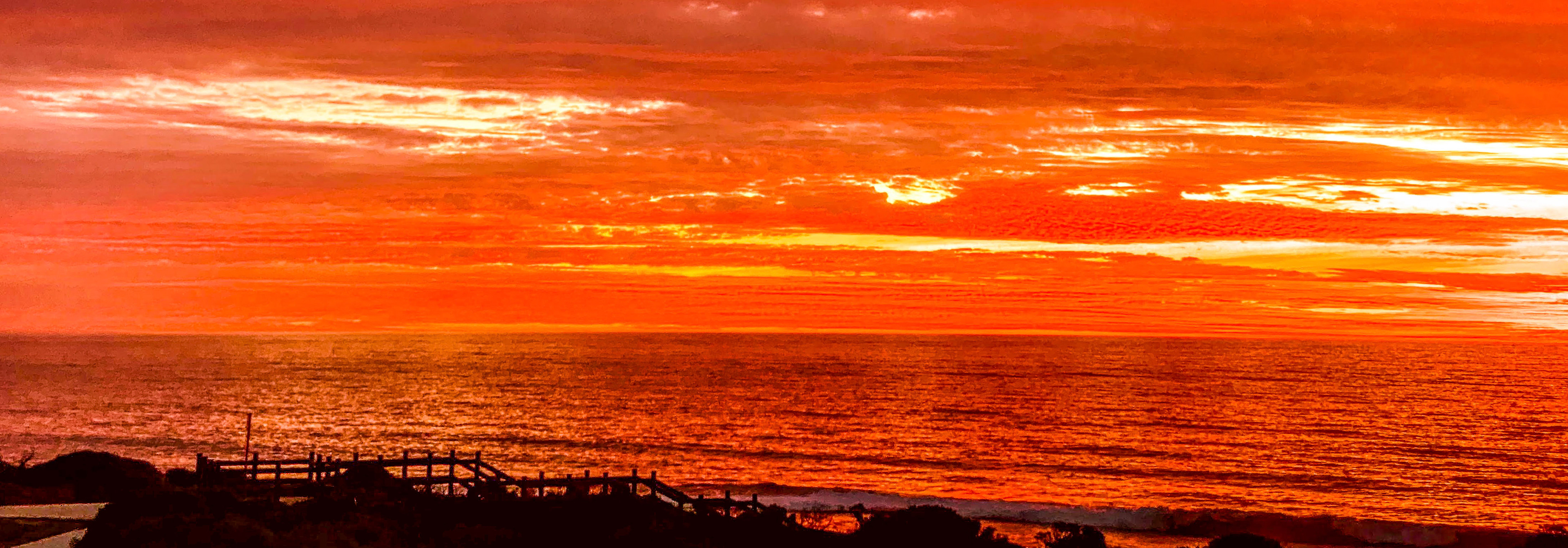 A dramatic sunset showcases vibrant orange and red hues in the sky, illuminating clouds over the serene ocean.
