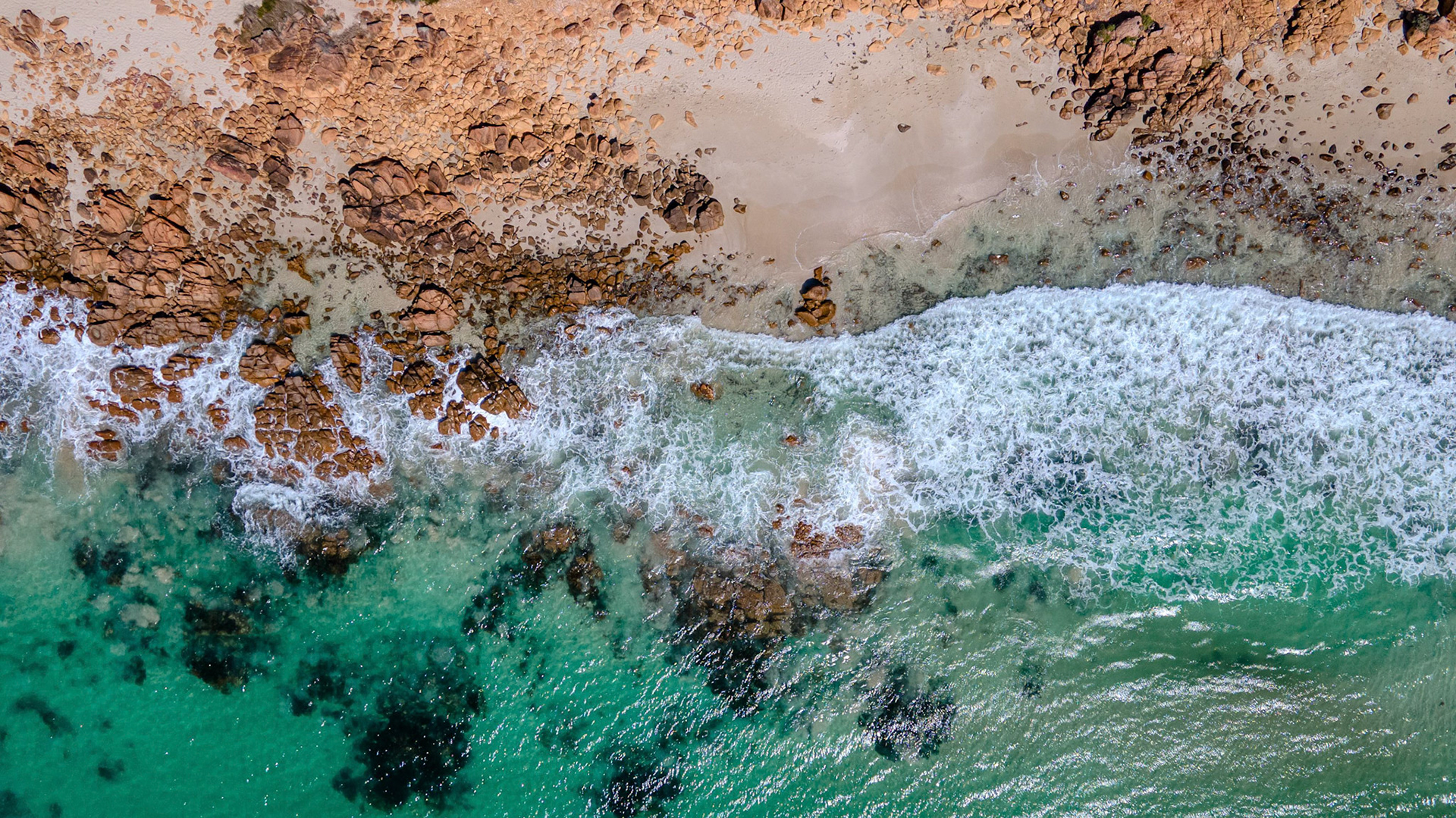Captivating aerial photograph showcasing ocean waves rhythmically crashing against a rugged, rocky shoreline. The image captures the stunning contrast between the white foam of the waves and the earthy tones of the rocks, alongside the vibrant turquoise of the clear ocean water. The natural patterns formed by the waves and rocks create a visually intriguing composition, ideal for nature-themed projects or backgrounds.